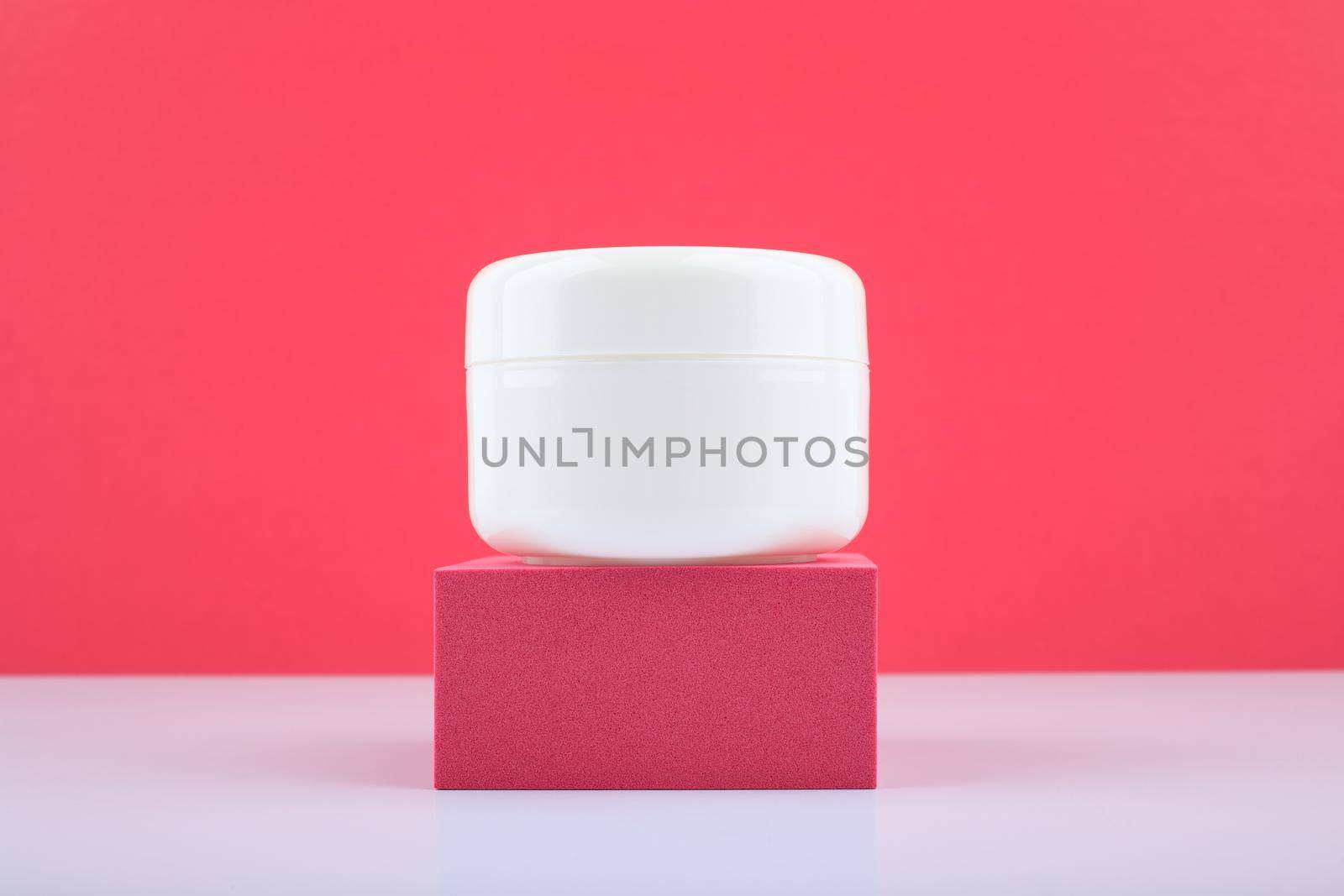 Simple composition with white glossy unbranded cream jar on pink podium against bright pink background. Concept of skin care and wellbeing