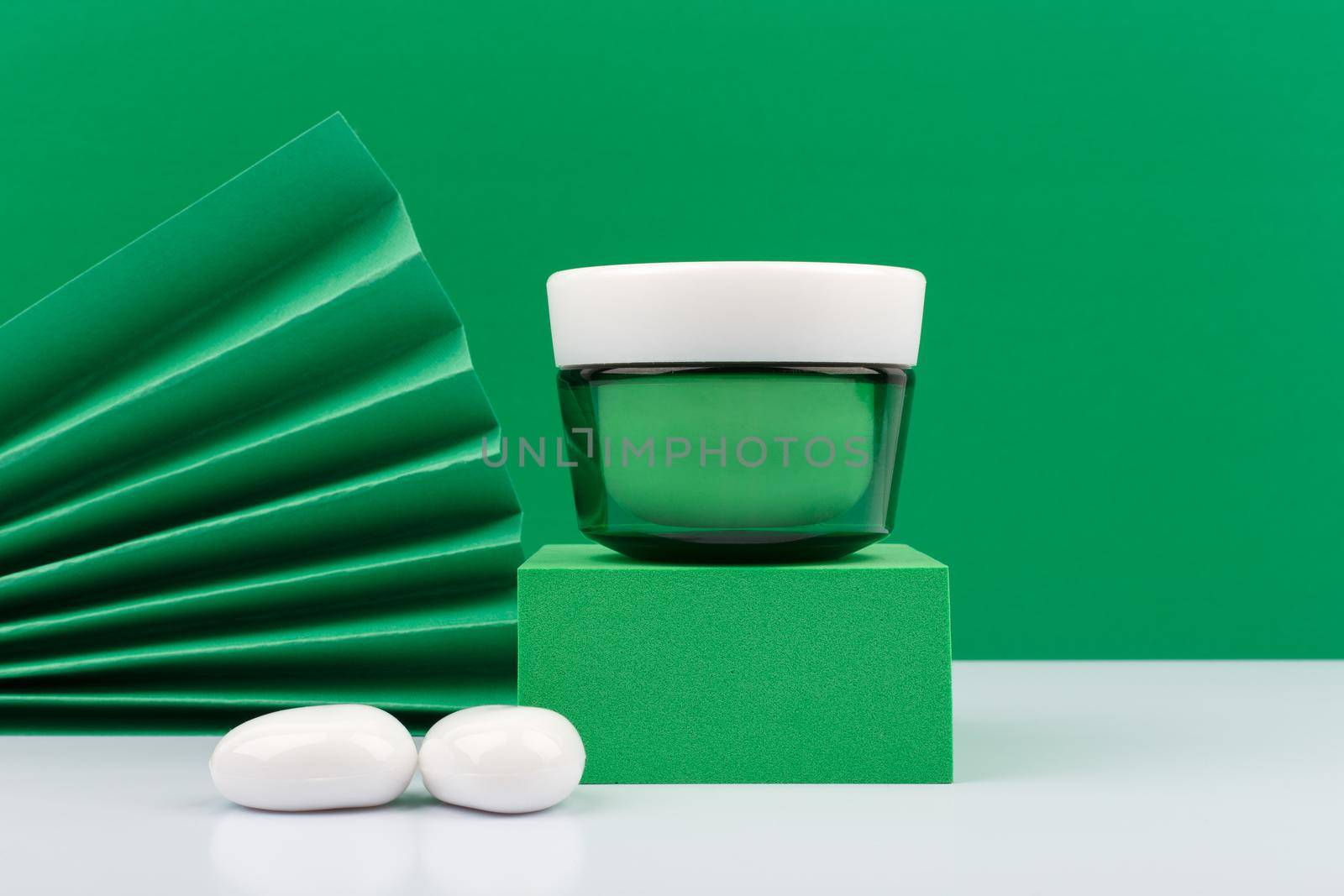 Still life with green glass cream jar on green podium against green background decorared with green waver and white stones