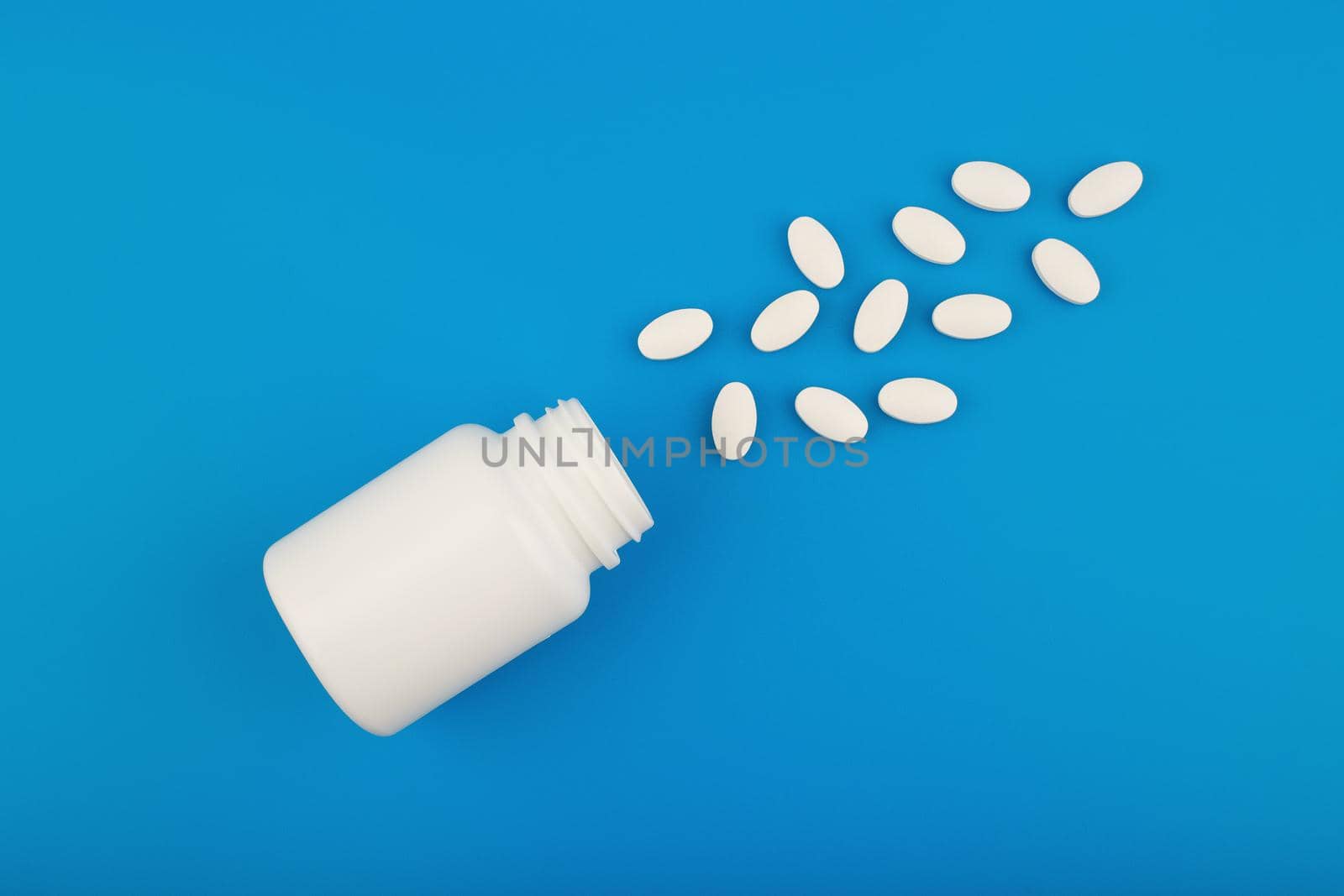White unbranded medication bottle with spilled oval pills on blue background. Concept of medical treatment, virus protection or vitamins