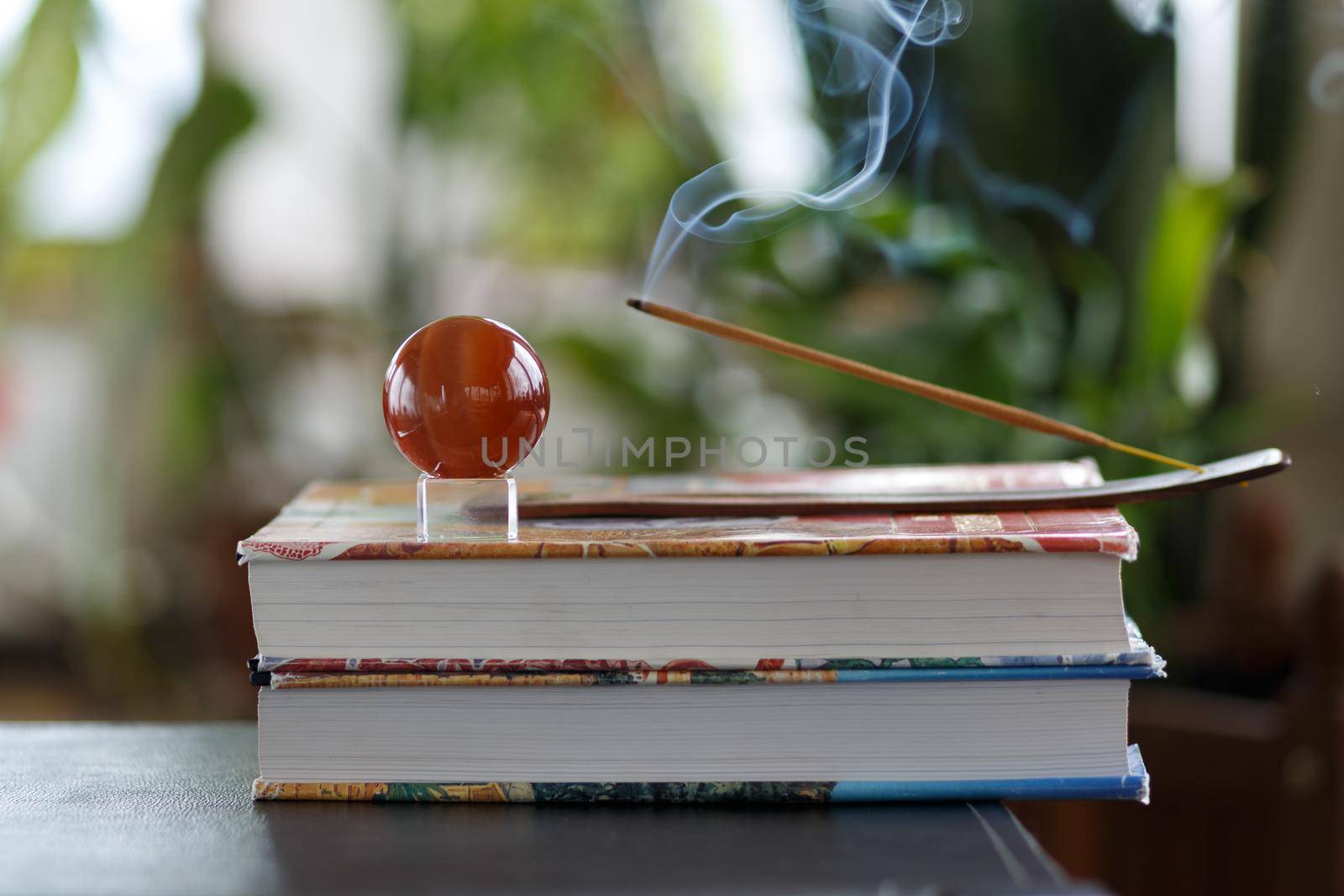 Selective focus, still life with burning incense stick and brown glass ball on a stack of magazines at home with blurred flowers at the background. Concept of staying home and relax or meditation