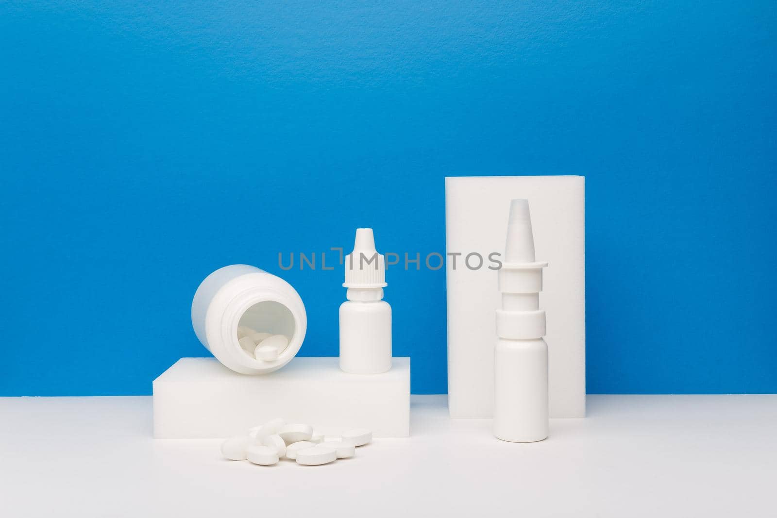 Still life with white medication bottle with spilled pills on white podium, nose spray and eye drops against blue background on white table