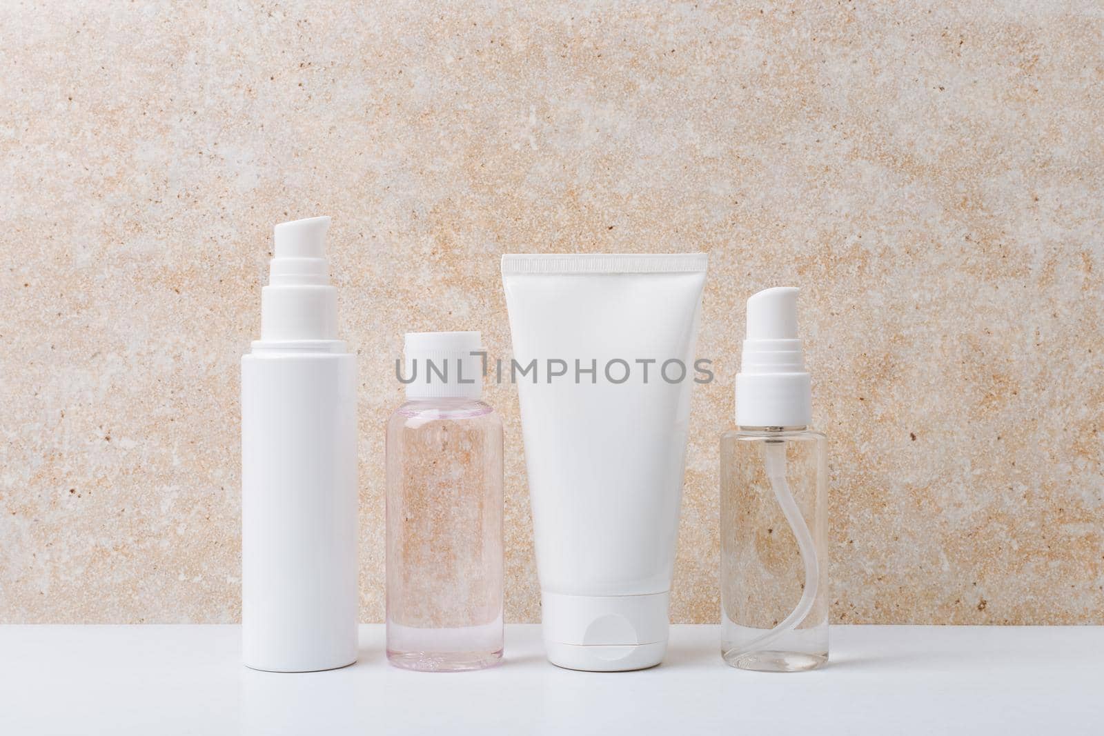Still life with a set of cosmetic products for daily skin care on white table against beige marble background. Face cream, cleaning foam, lotion and hands cream in unbranded bottles