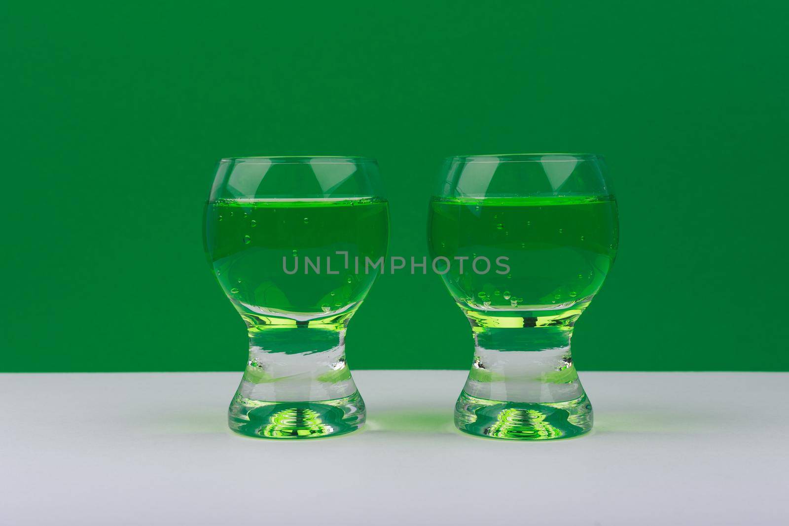 Two glass shots with green drinks on white table against green background by Senorina_Irina