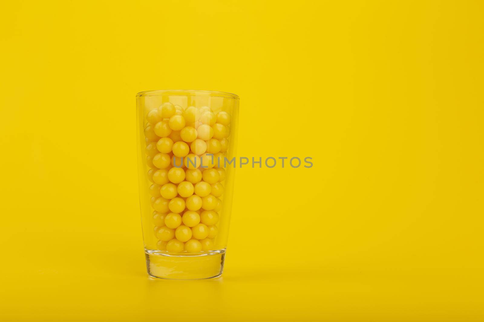 Transparent glass shot with yellow round vitamins on yellow background with space for text. The concept of vitamin C and vitamin D or healthy lifestyle