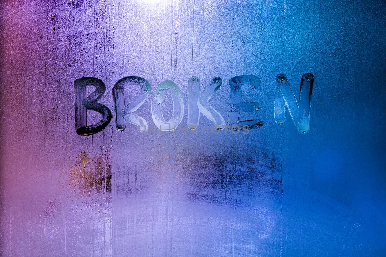 the word broken handwritten on wet window glass with selective focus and backgropund blur, neon blue color