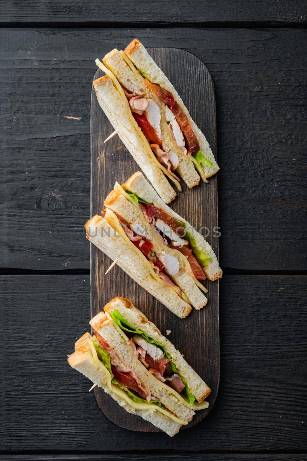 Homemade club sandwich made with turkey, bacon, ham, tomatoes, on black wooden table, top view