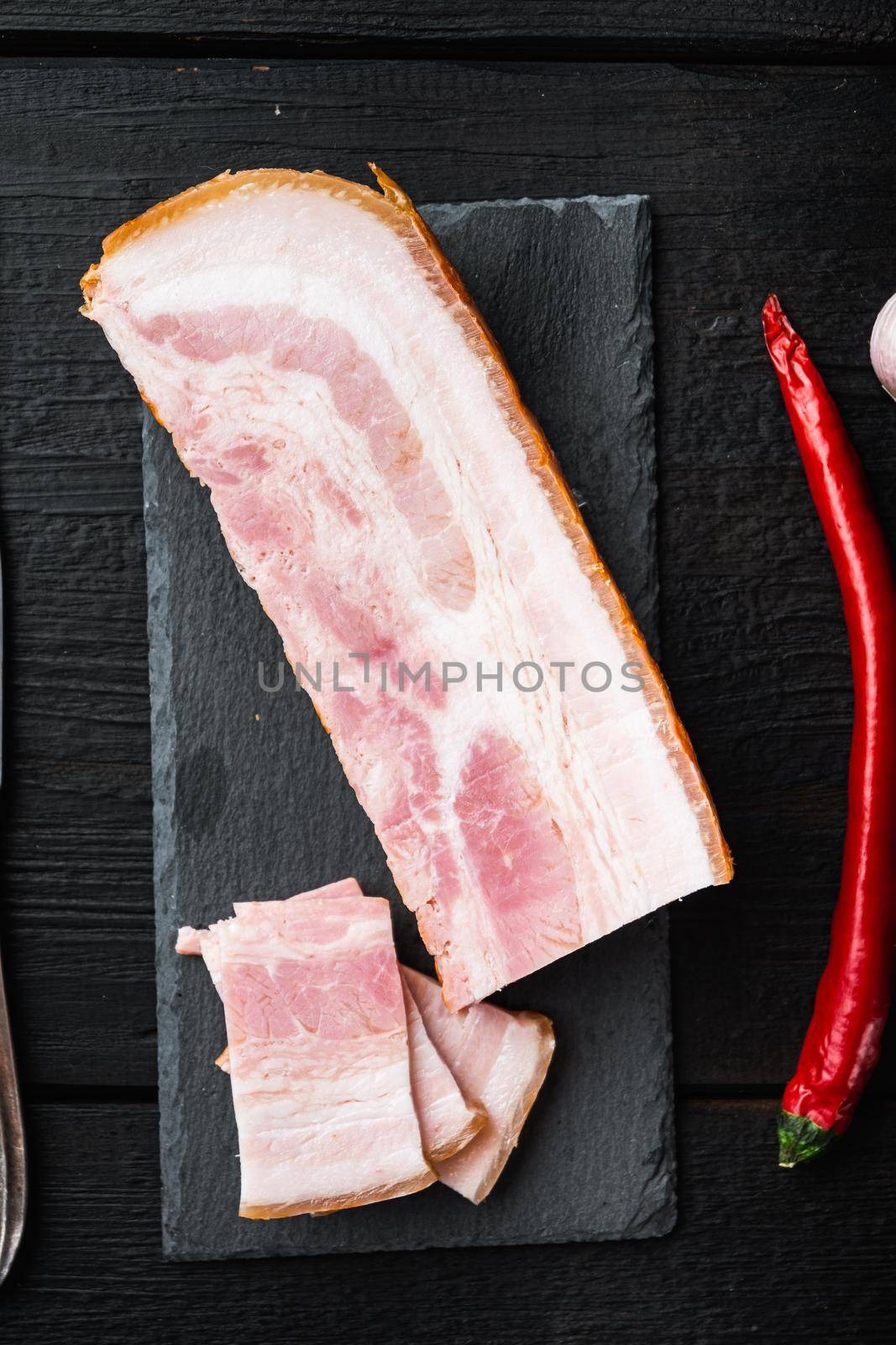 Smoked bacon, whole slab with herbs on black wooden table, top view.