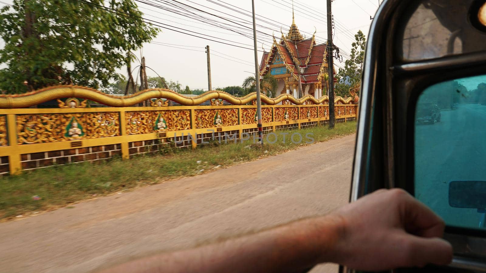 Travel by car in Thailand. View of the road. Urban landscape of Trat province. I move around in trunk of a pickup truck. Passing cars, motorcyclists. There are houses, trees, billboards and a temple.