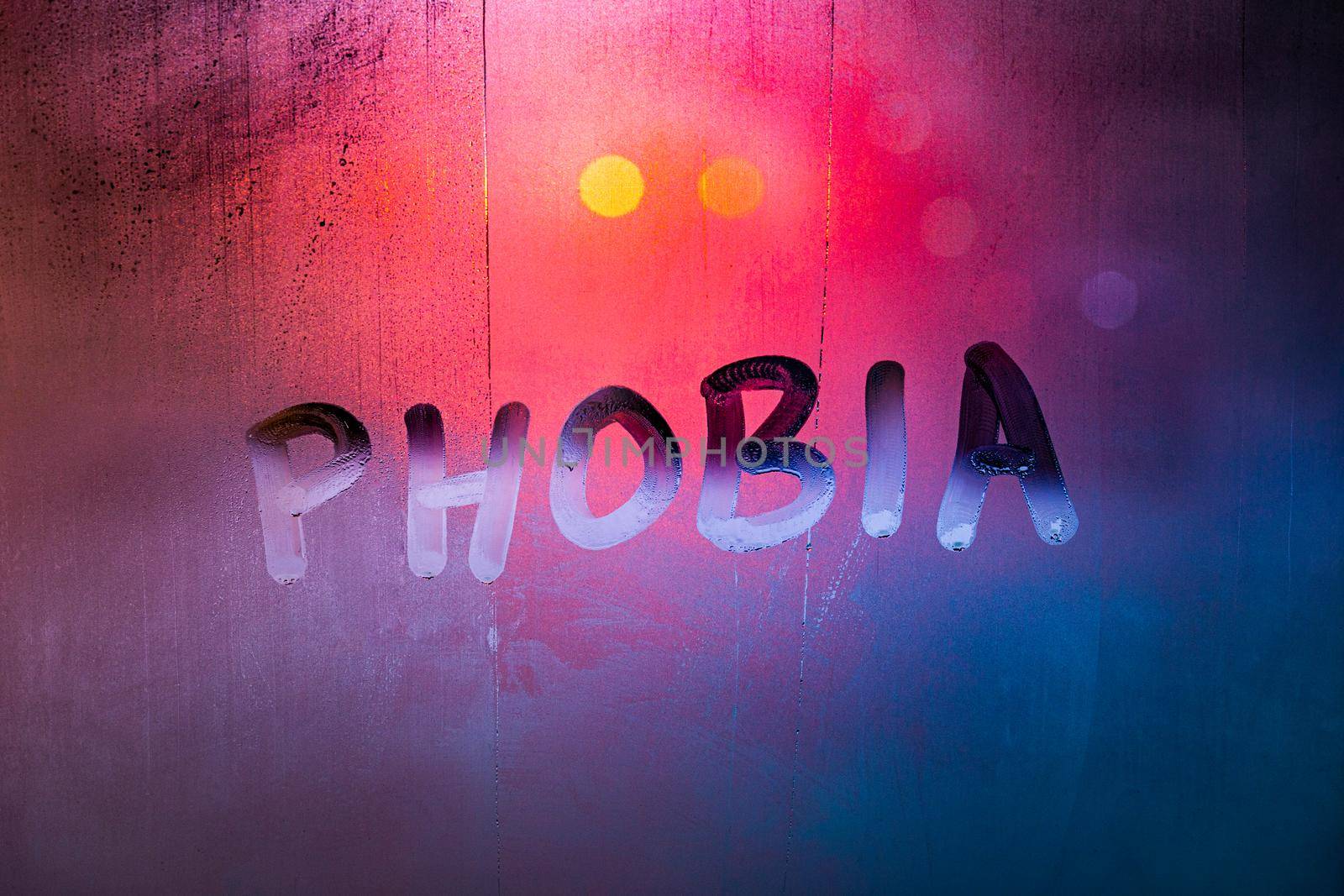 the word phobia handwritten on wet foggy window glass surface with red blue background lights.