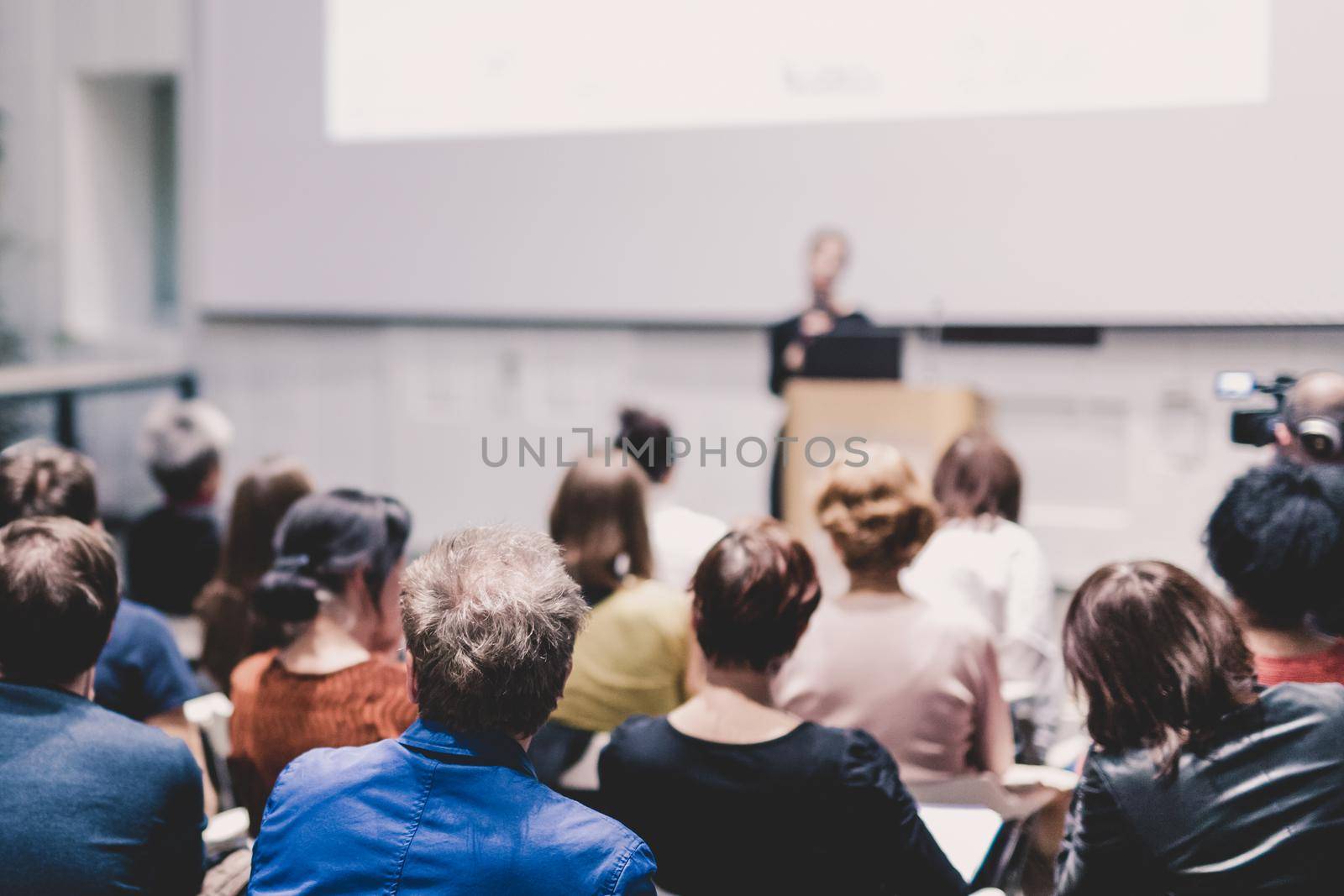 Business and entrepreneurship symposium. Female speaker giving a talk at business meeting. Audience in conference hall. Rear view of unrecognized participant in audience. Copy space on whitescreen.