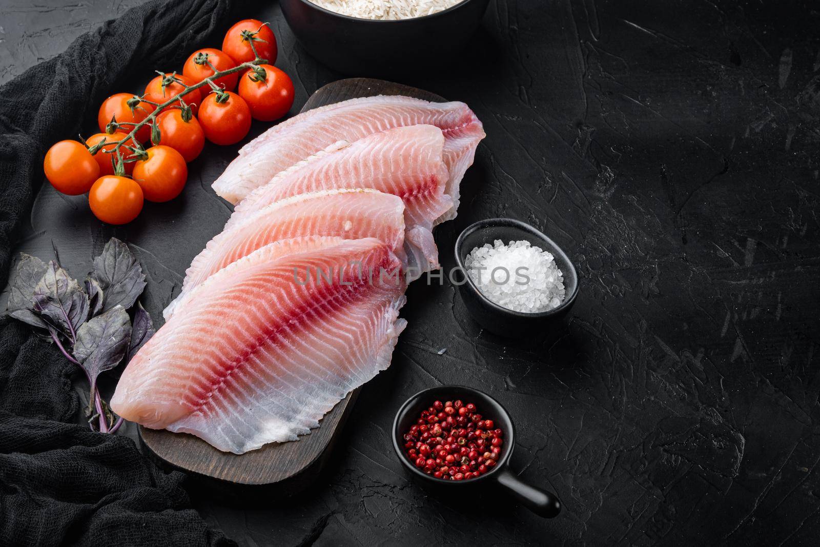 Raw white fish tilapia, with basmati rice and cherry tomatoes ingredients, on black background