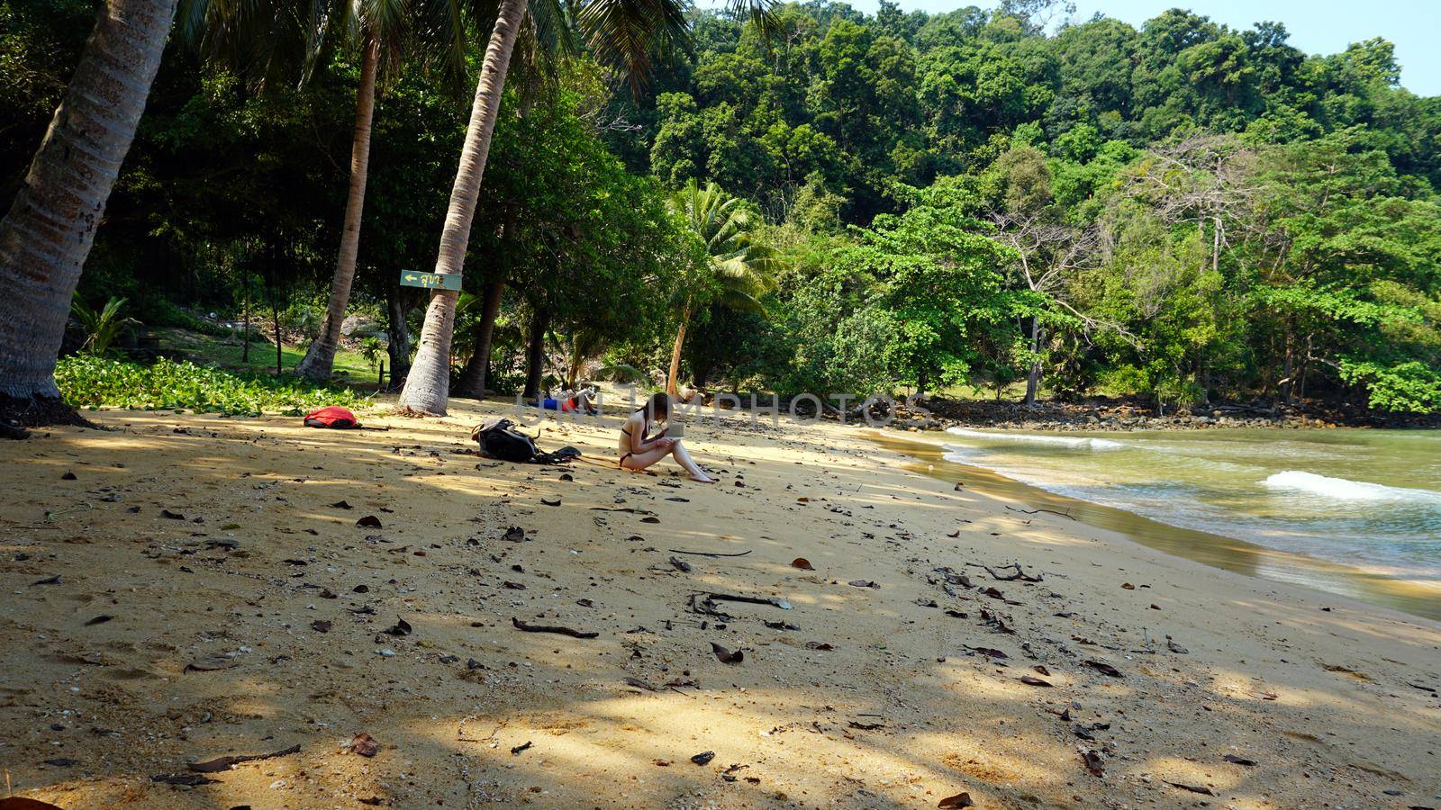 A girl in a bathing suit reads a book on the beach. Green island, lots of trees and palm trees. Small waves from the green water. In the distance, a guy lies under the shade of trees. Thailand.