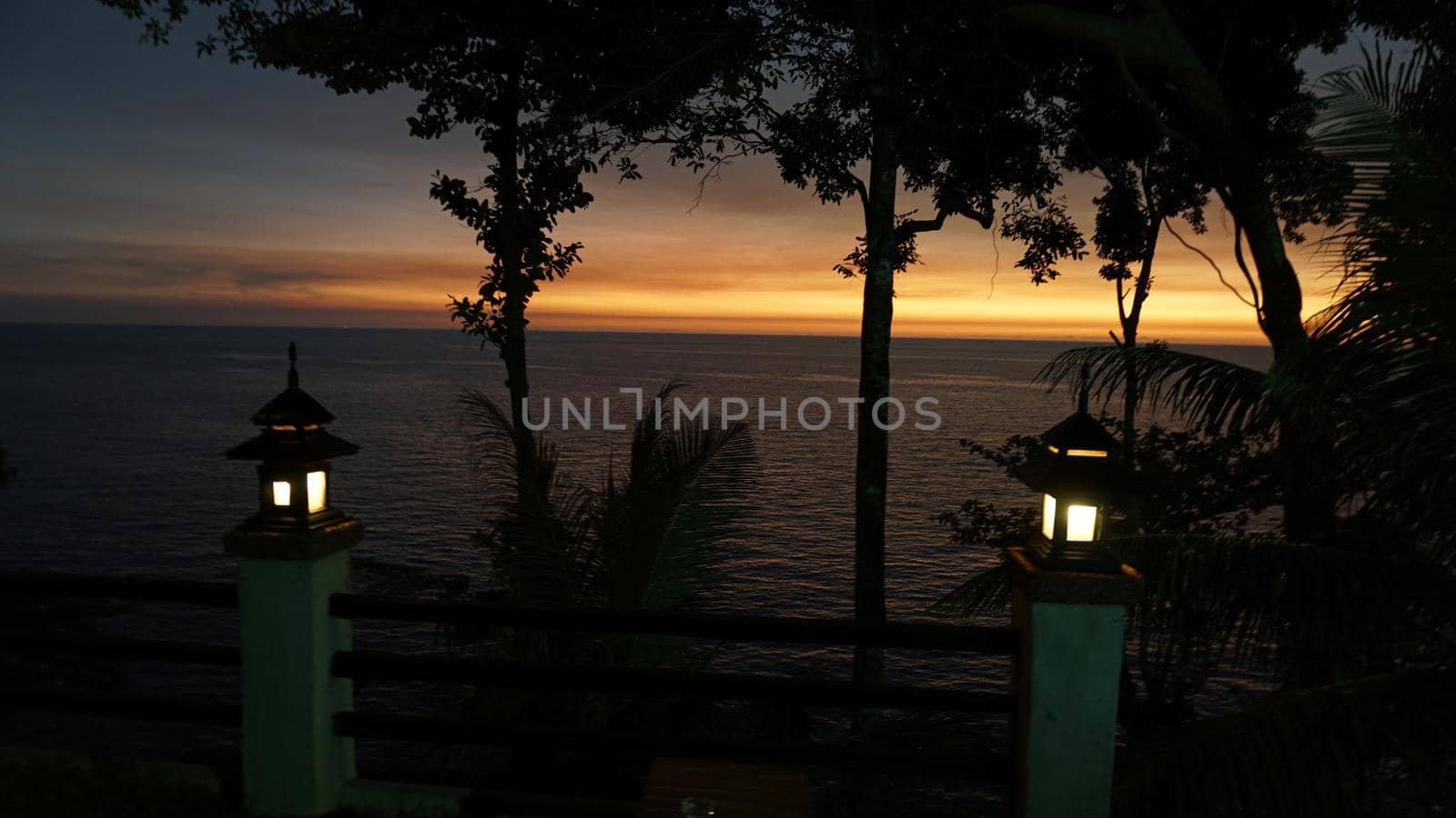 The sunset with views of the sea and palm trees. The sun goes below the horizon. An orange ray of sunlight is reflected on the water. Palm trees stand near the sea. Small clouds float across the sky.