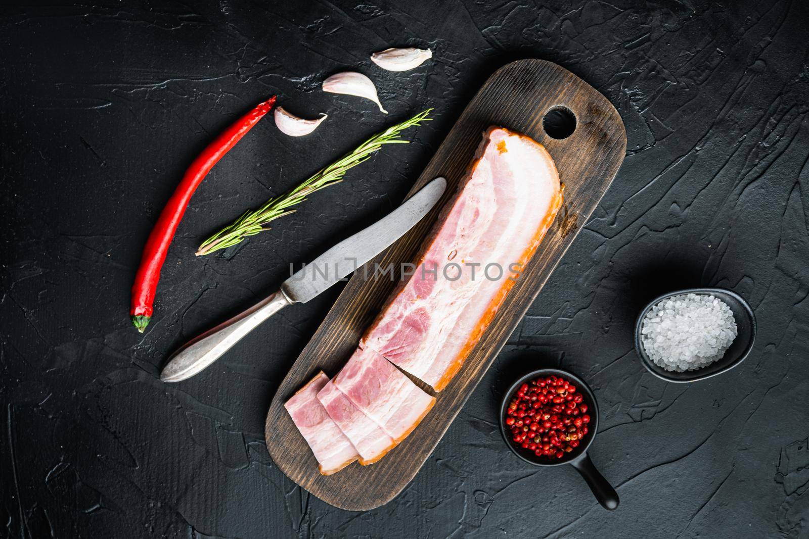 Bacon pancetta cut and sliced with herbs on black background, top view.