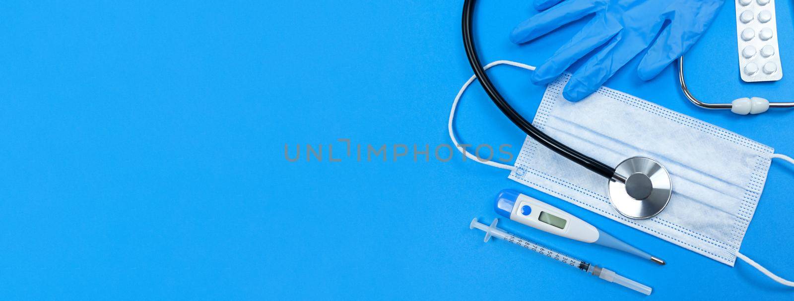 Stethoscope, face mask, medical syringe, thermometer, pills blister and medical gloves on a blue background. Flat lay banner with copy space.