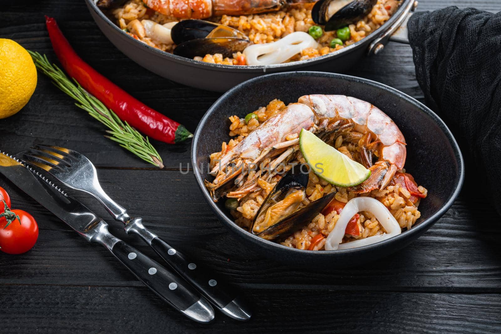 Traditional spanish seafood paella in pan and bowl with rice, peas, shrimps, mussels and squid on black wooden planks, food photo.