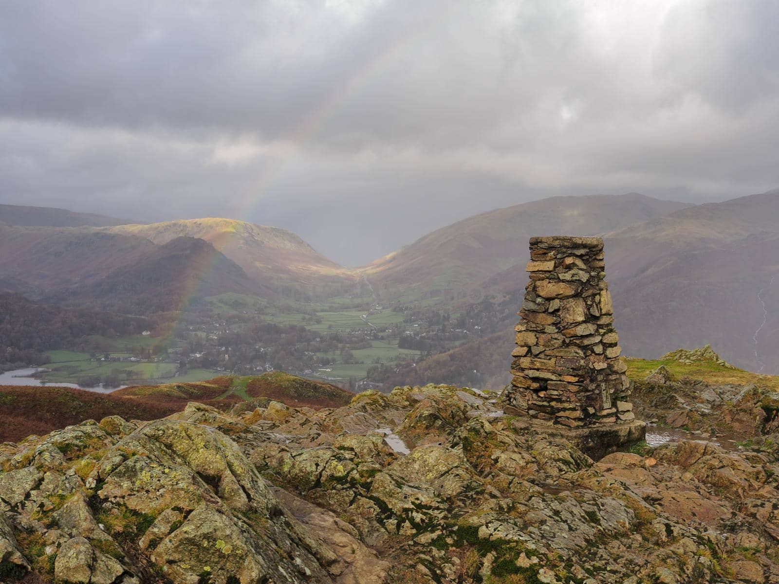 Triangulation point at the top of Loughrigg Fell overlooking Grasmere with a rainbow arcing across the fells under a stormy sky, Lake District, UK