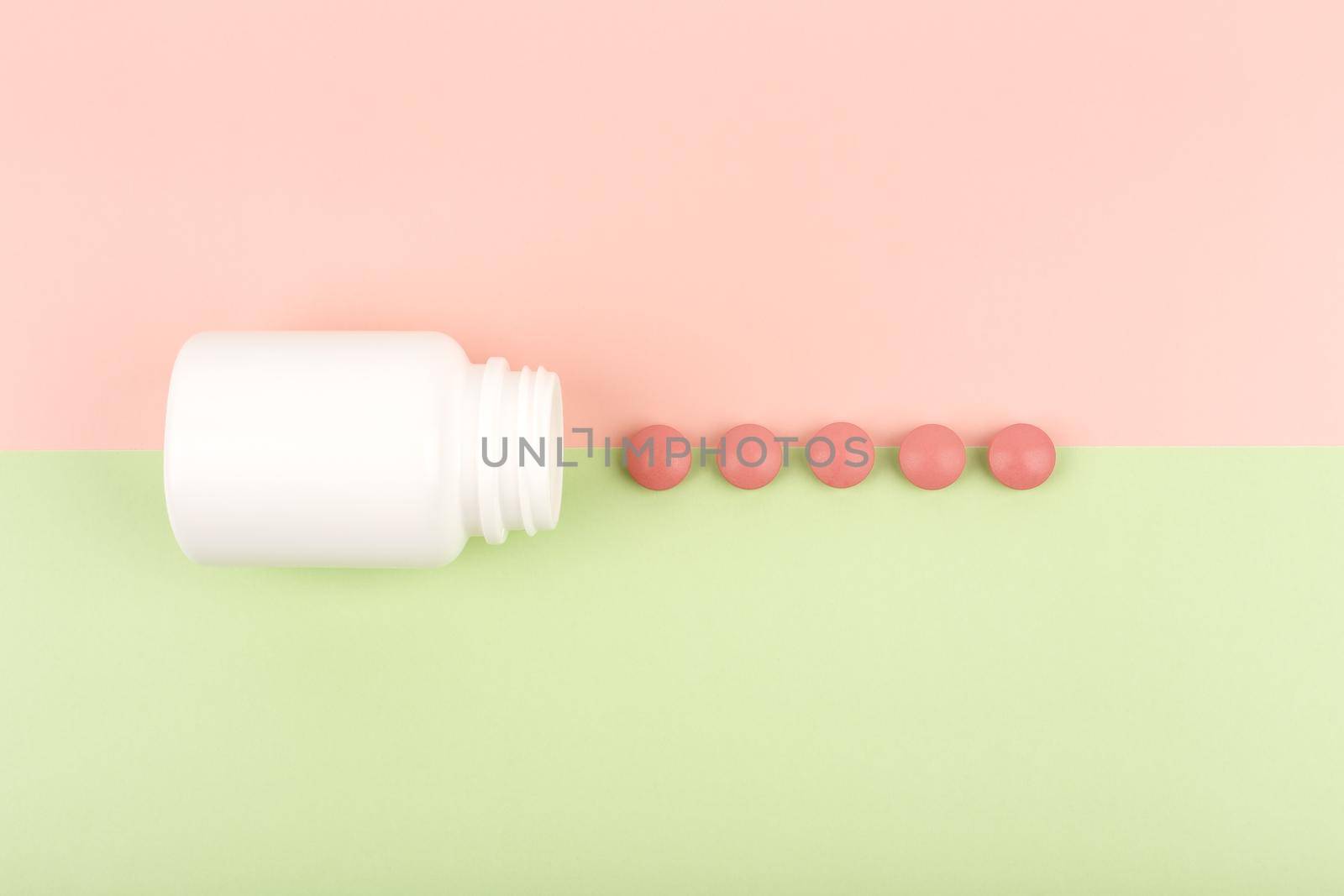 Top view of an opened white medication bottle with spilled red round pills on green and pink background. Concept of pharmacy and medications