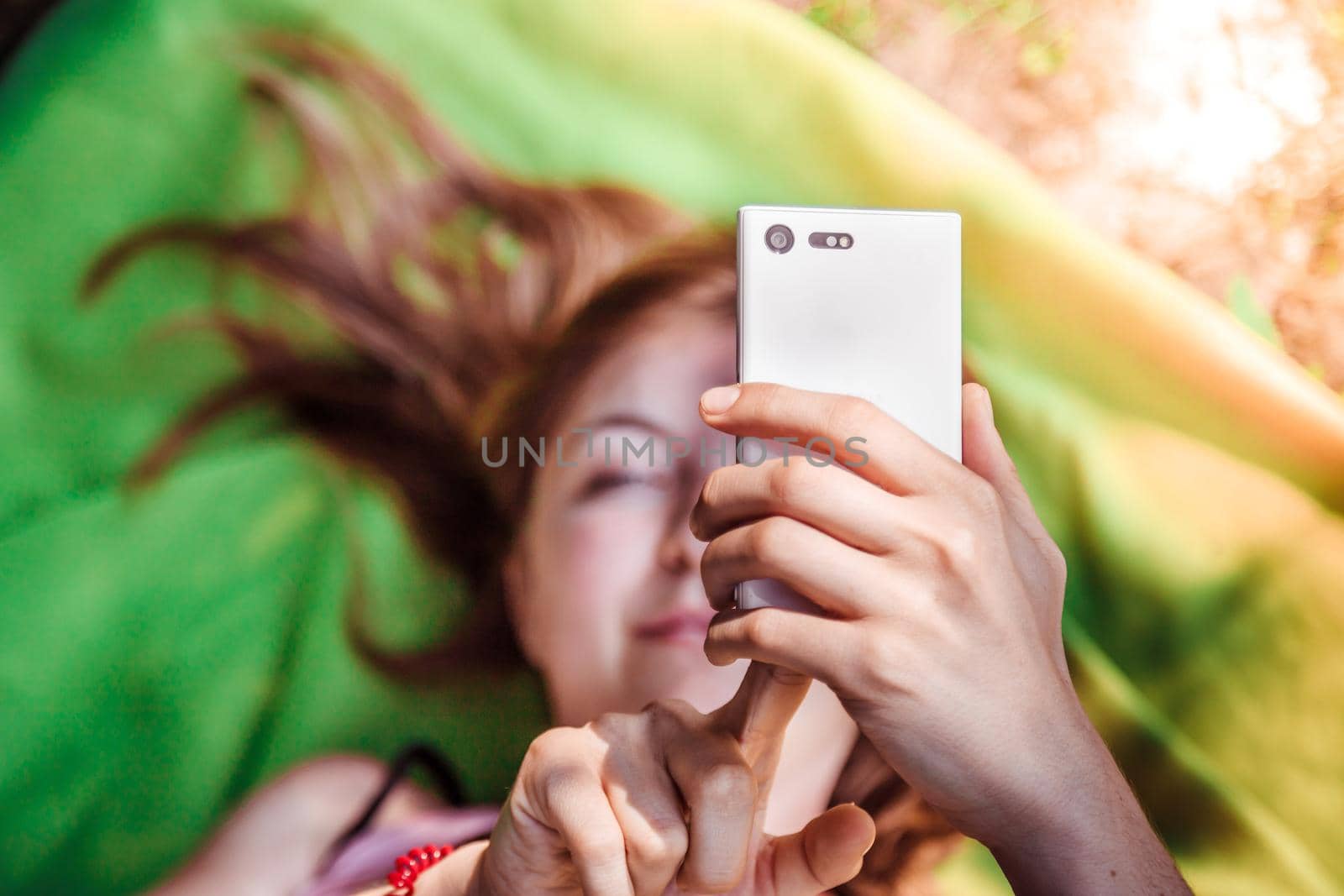 Beautiful girl is using her smartphone lying on a blanket in the park, above view