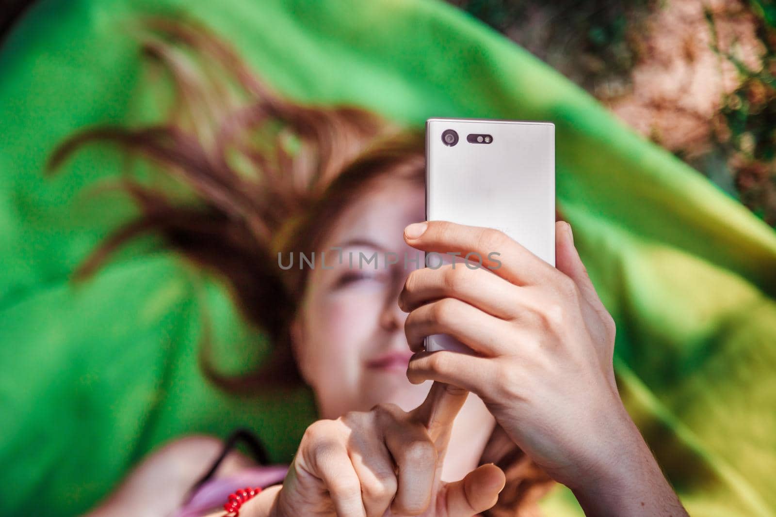 Beautiful girl is using her smartphone lying on a blanket in the park, above view