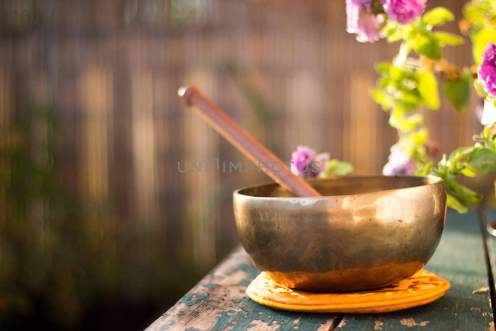 Metal singing bowl on a rustic green, wooden table outdoors. Flowers in the colourful, blurry background