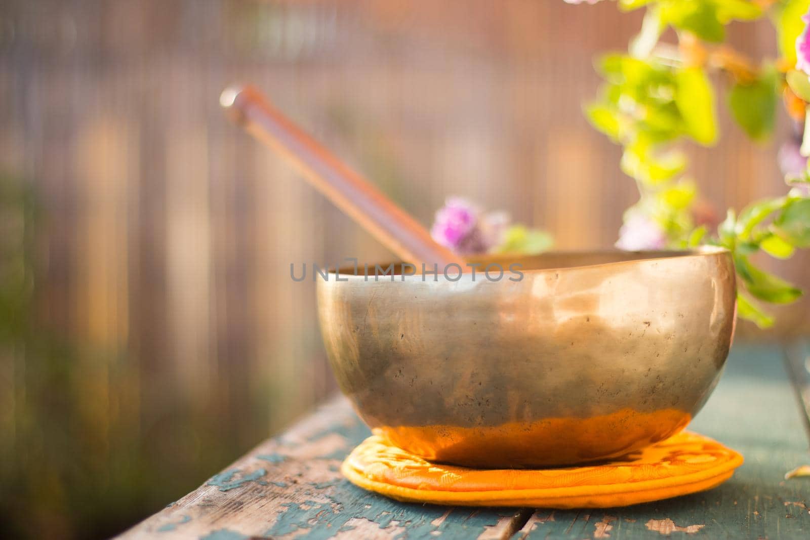 Metal singing bowl on a rustic green, wooden table outdoors. Flowers in the colourful, blurry background