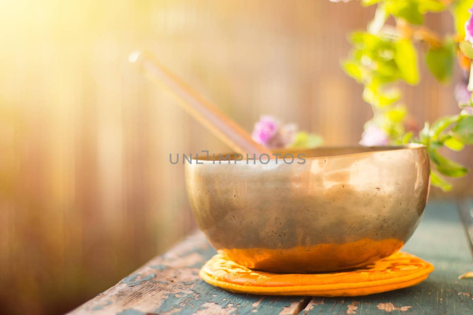 Metal singing bowl on a rustic green, wooden table outdoors. Flowers in the colourful, blurry background