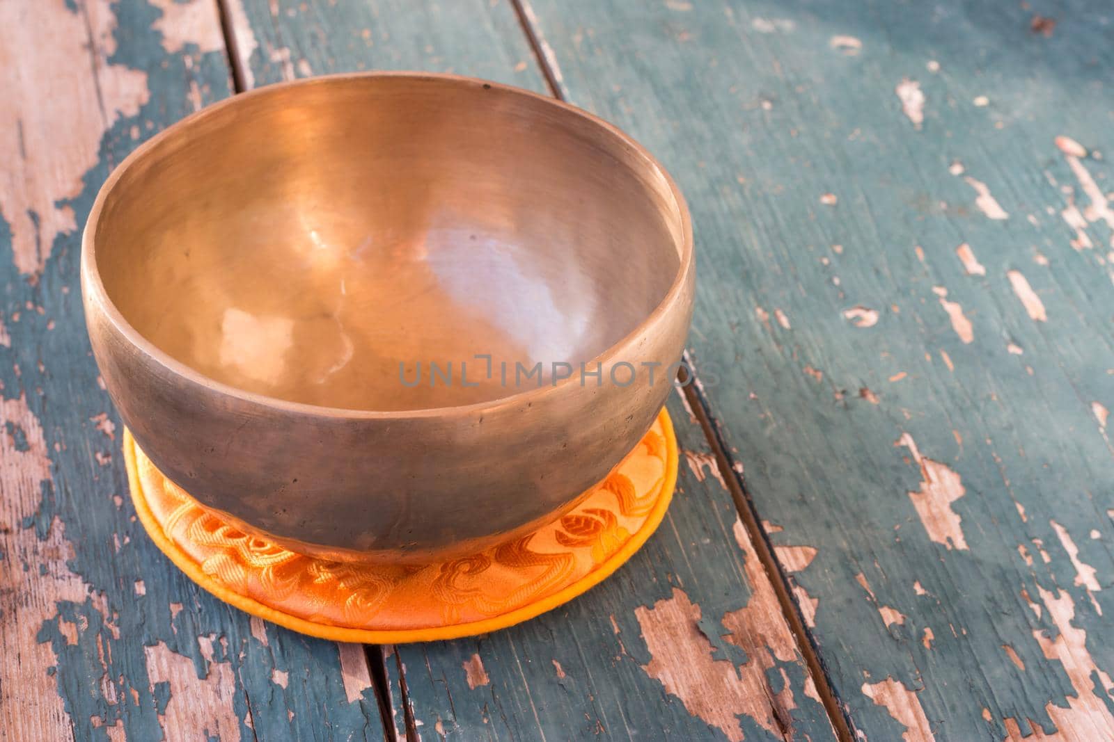 Metal singing bowl on a rustic green, wooden table outdoors. Flowers in the colourful, blurry background