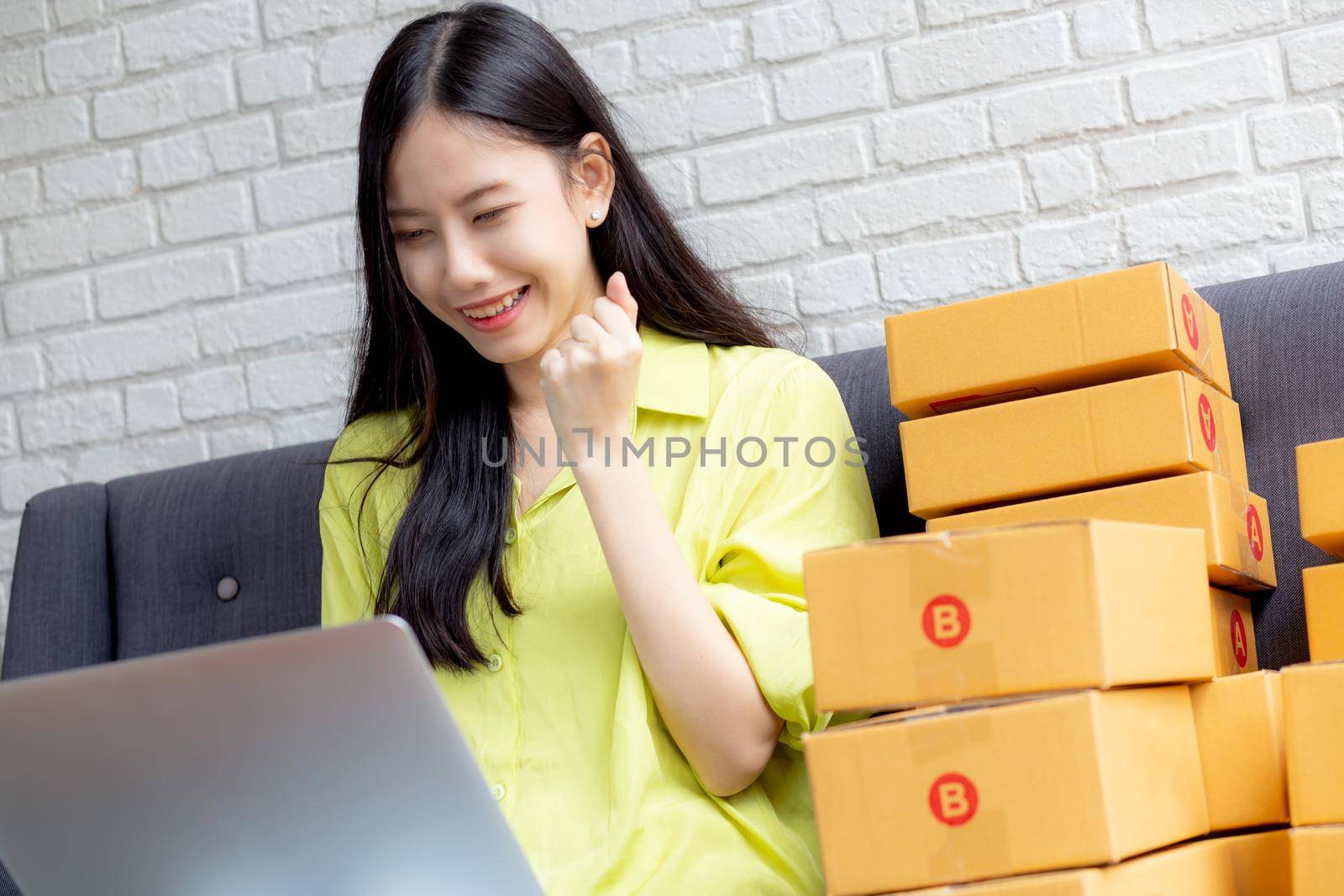 Young asian woman is merchant using laptop computer on sofa checking order of customer with purchase online shopping, business SME or startup, entrepreneur or seller and service delivery product.