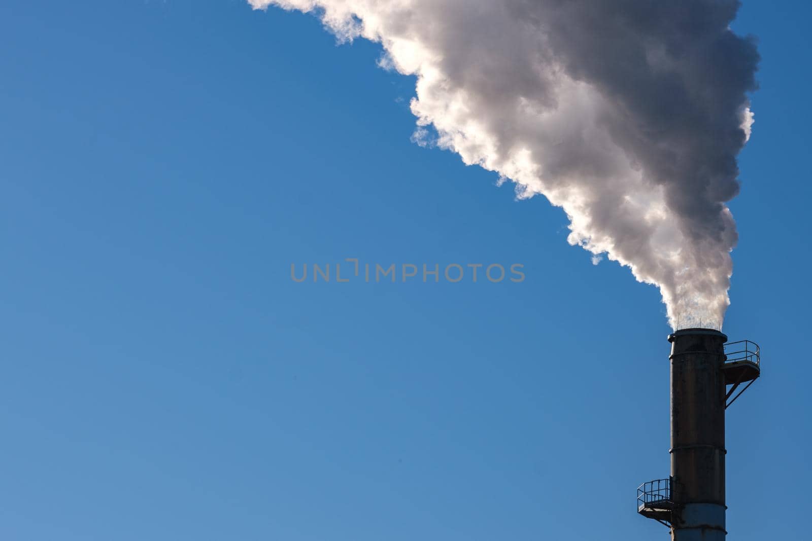 Thick smoke billows into a clear blue sky from the top of a tall smokestack.