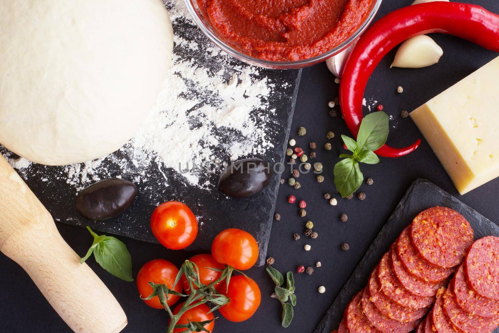 Raw dough for pizza with ingredients and spices on black table background