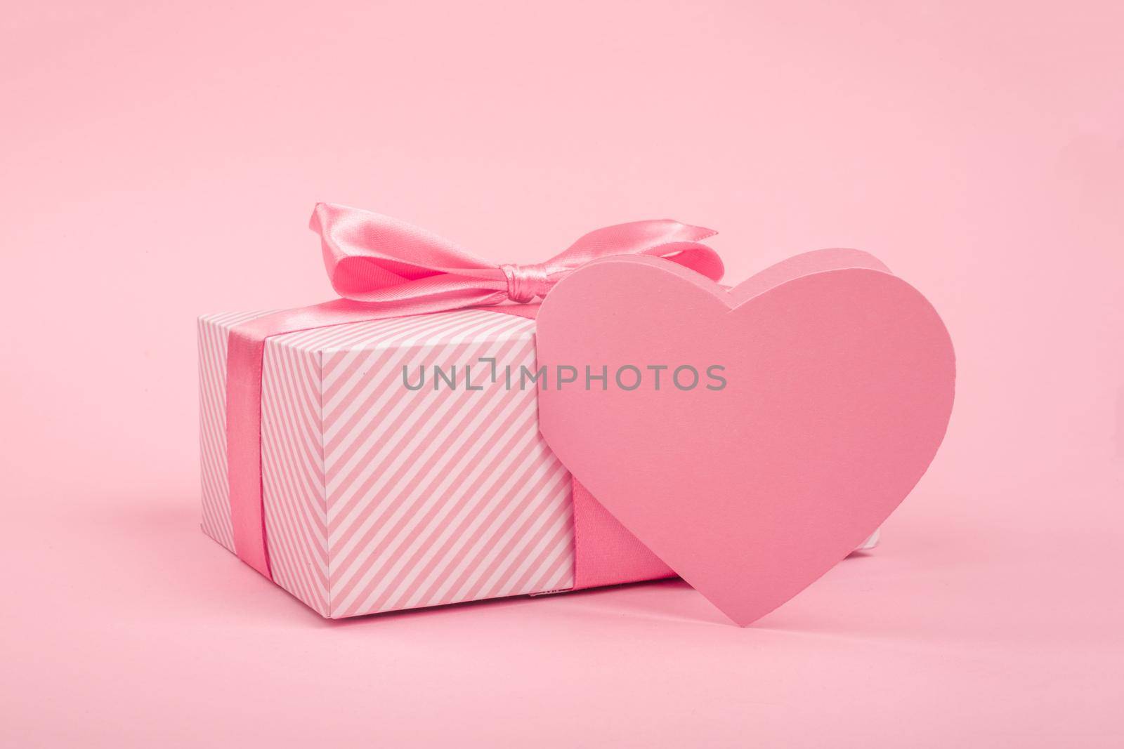 Valentine Day gift in a box wrapped in striped paper and tied with silk ribbon bow and heart shapes greeting card on pink background with copy space for text