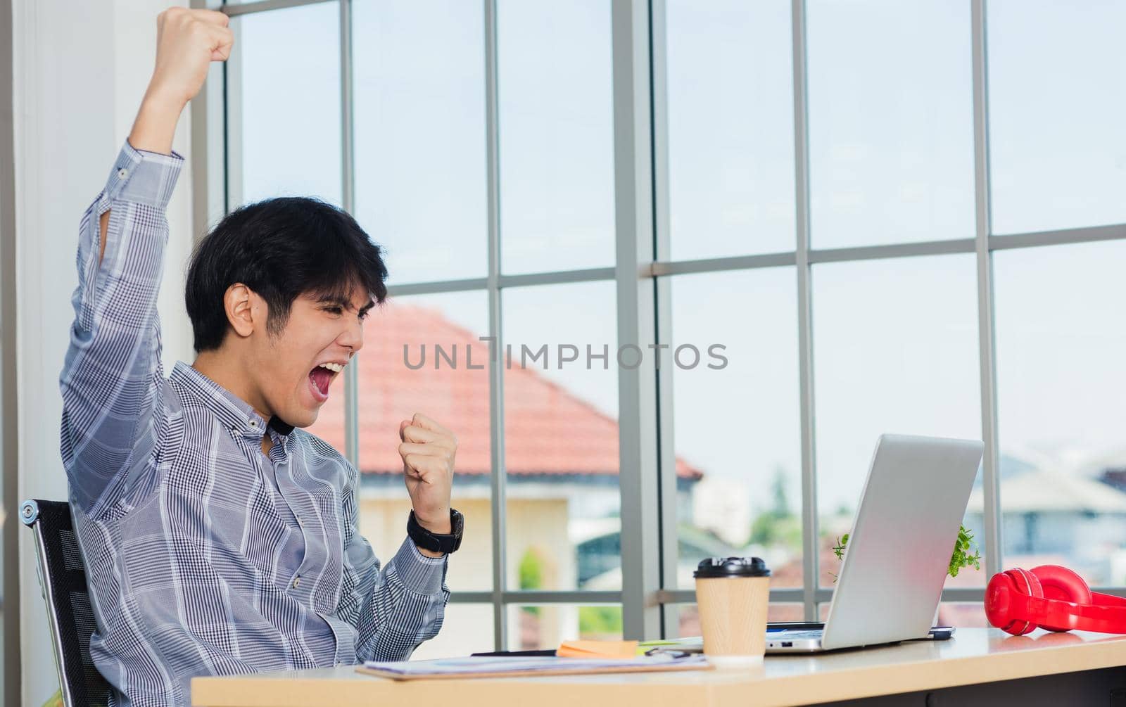 Yes! Happy excited Asian man raising his arm up to celebrate celebrating success. Young businessman using laptop computer at office desk he glad to receive good winner profits from the job