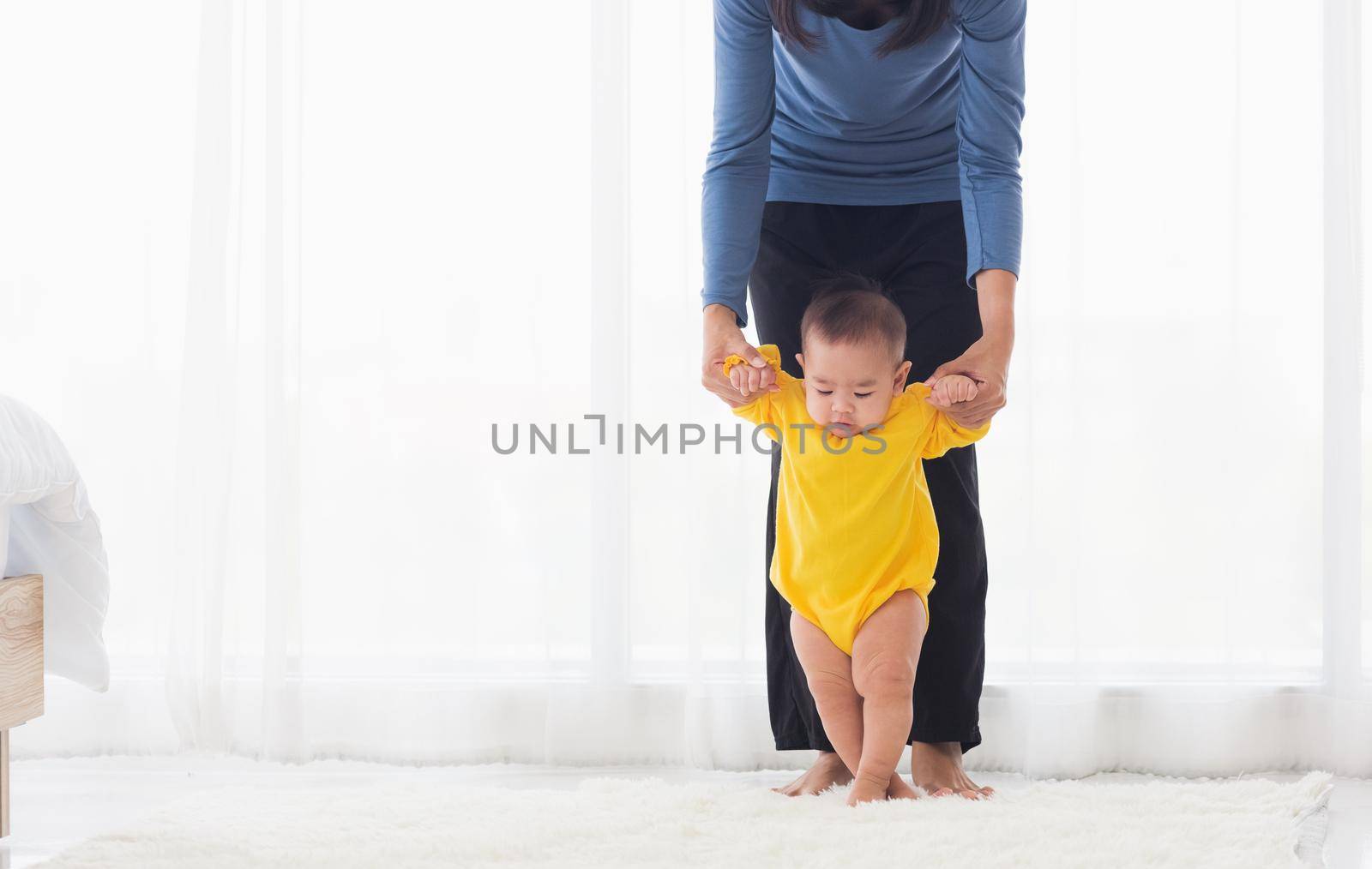 Asian little baby girl taking first steps learning to walk with mom help support the cute unstable walking toddler at home in bedroom. Happy family first steps parenthood concept