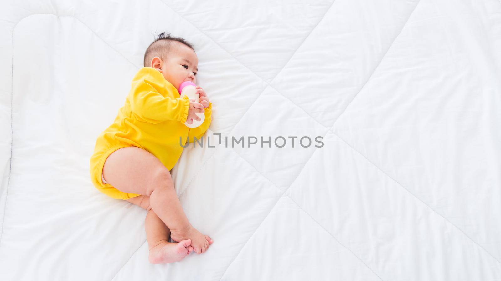 Asian beautiful little baby girl wearing a yellow dress eat milk sleeping feeding lying on the white bed, infant holding a bottle of milk, baby food concept