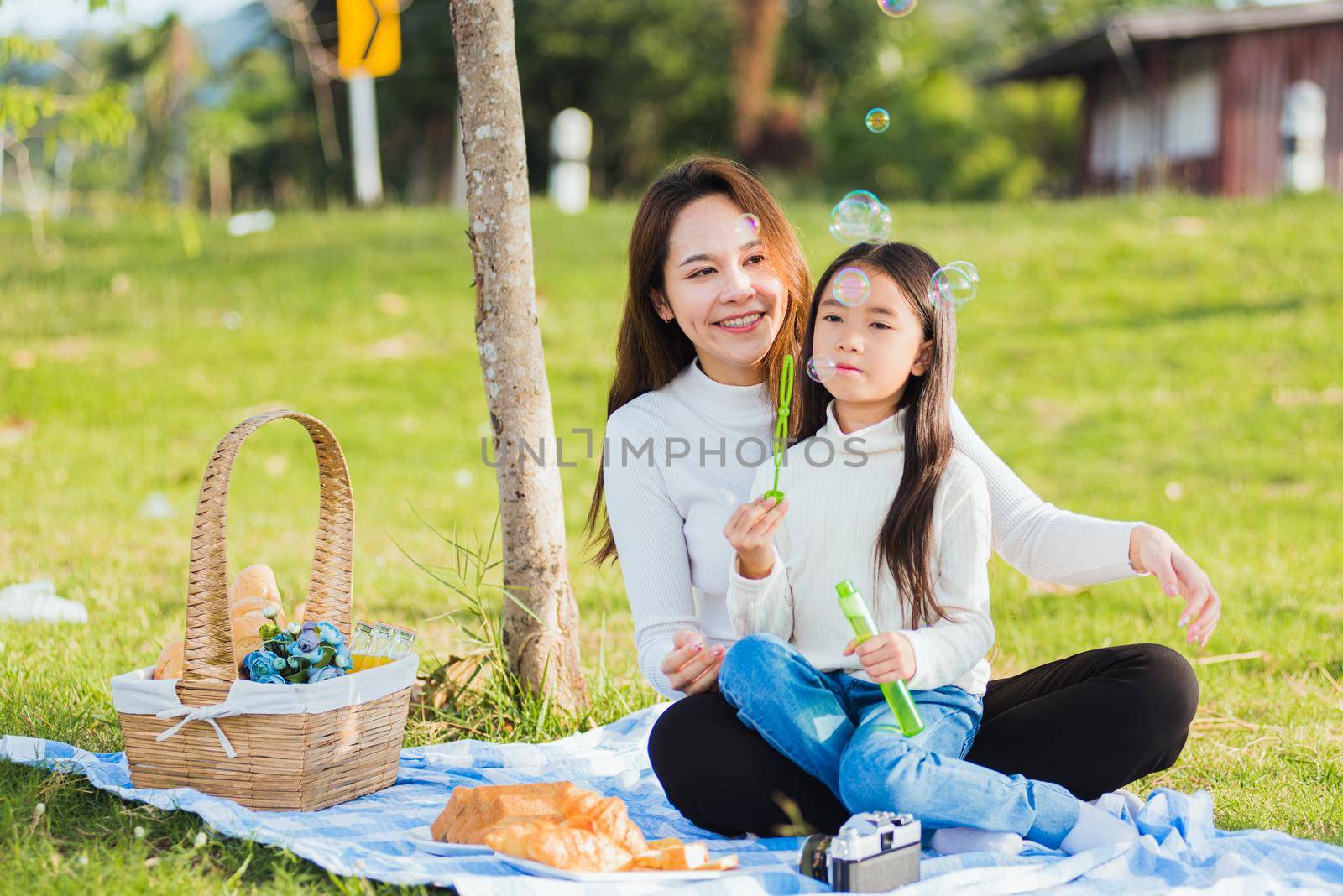 Happy Asian mother and little girl daughter child having fun and enjoying outdoor together sitting on the grass blowing soap bubbles during a picnic in the garden park on a sunny day family time