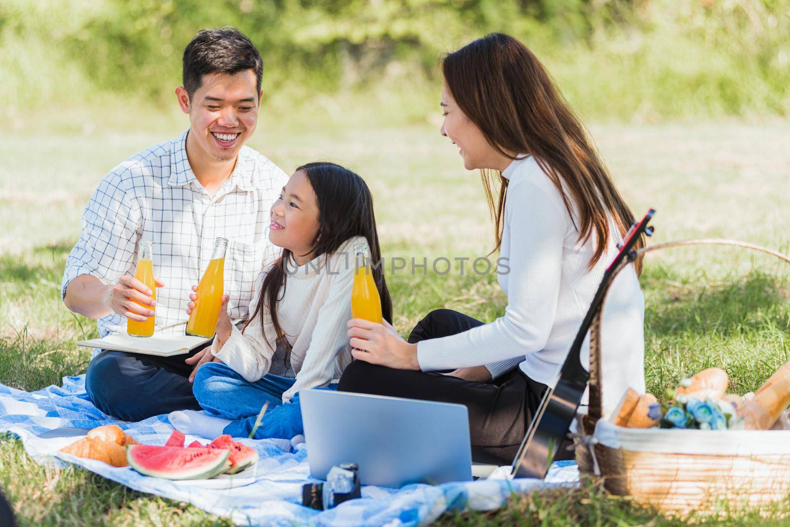 Happy Asian young family father, mother and child little girl having fun and enjoying outdoor sitting on picnic blanket drinking orange juice from glass bottle, Summer resting at a nature garden park