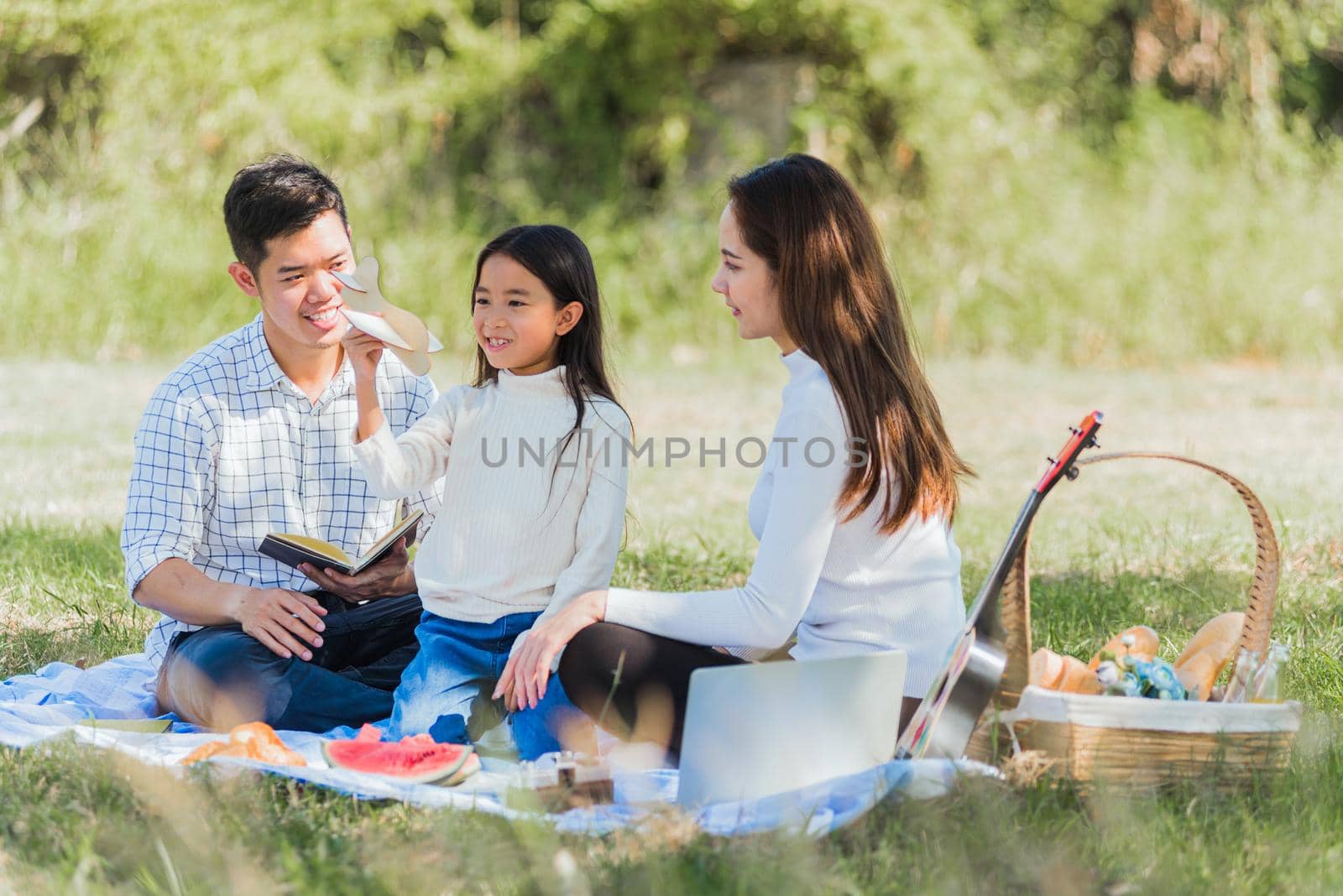 Happy Asian young family father, mother and child little girl having fun and enjoying outdoor sitting on picnic blanket playing toy aircraft at summer garden park