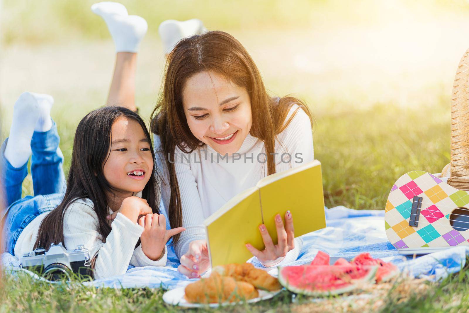 Happy Asian young family mother and child little girl having fun and enjoying outdoor laying on picnic blanket reading book at summer garden spring park, Family relaxation concept