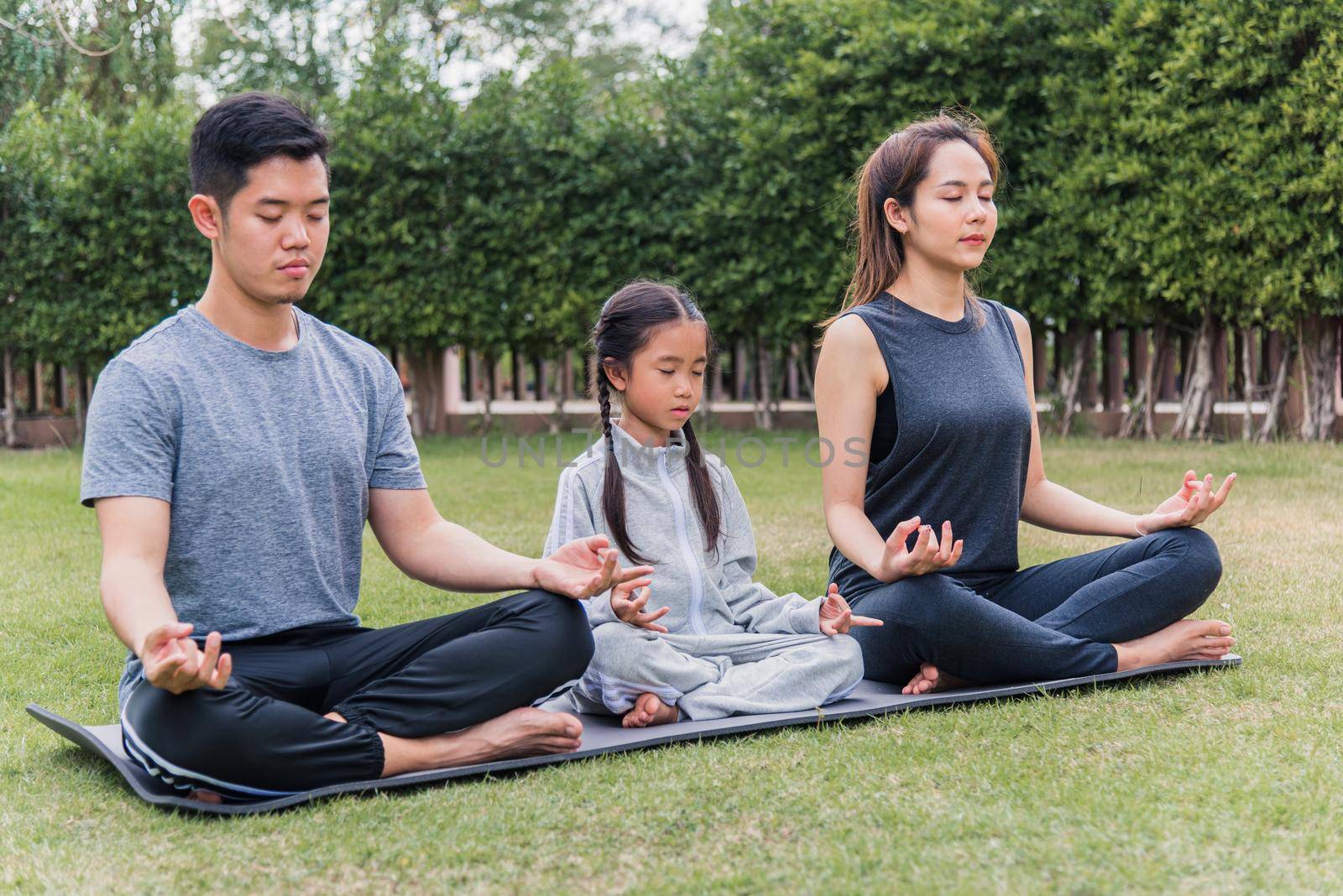 Asian young mother, father practicing doing yoga exercises with child daughter outdoors in meditate pose together in nature a field garden park, family kid sport and exercises for healthy lifestyle