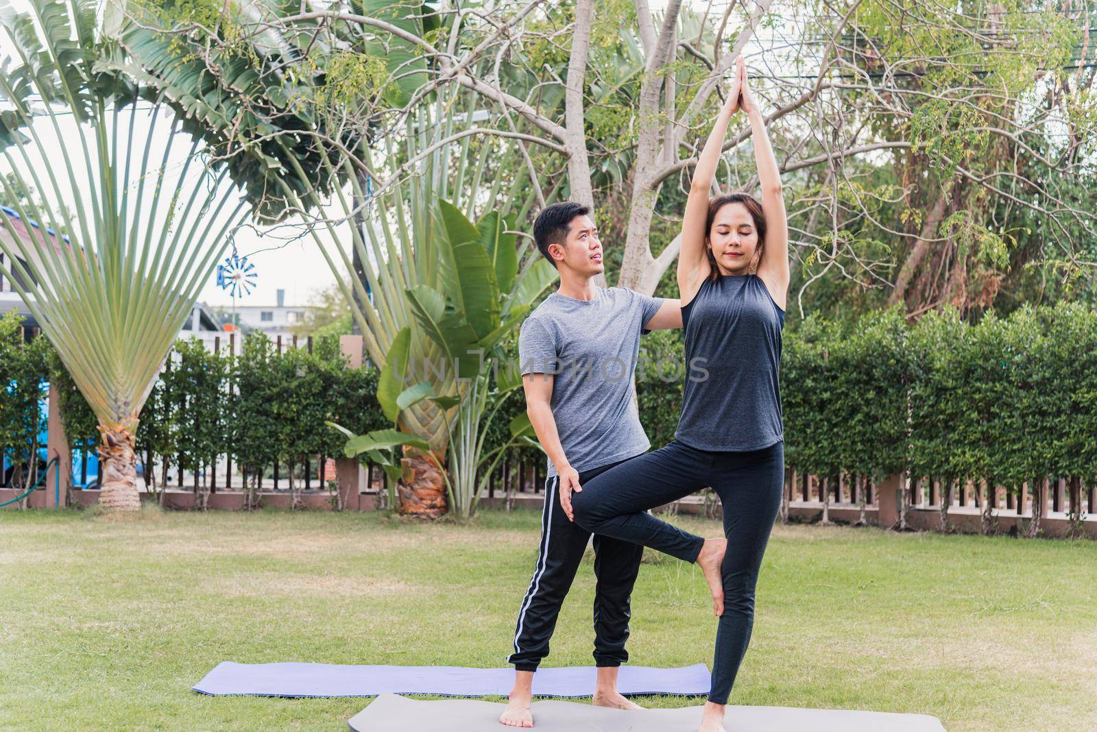 Asian man and woman training yoga outdoors in meditate pose stand on green grass. Young couple practicing doing stretching in nature a field garden park together. Meditation health care concept