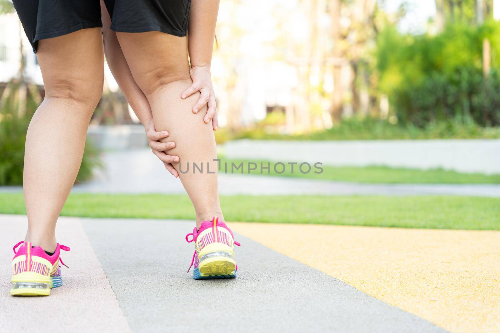Female fatty runner athlete leg injury and pain. Hands grab painful leg while running in the park. by mikesaran