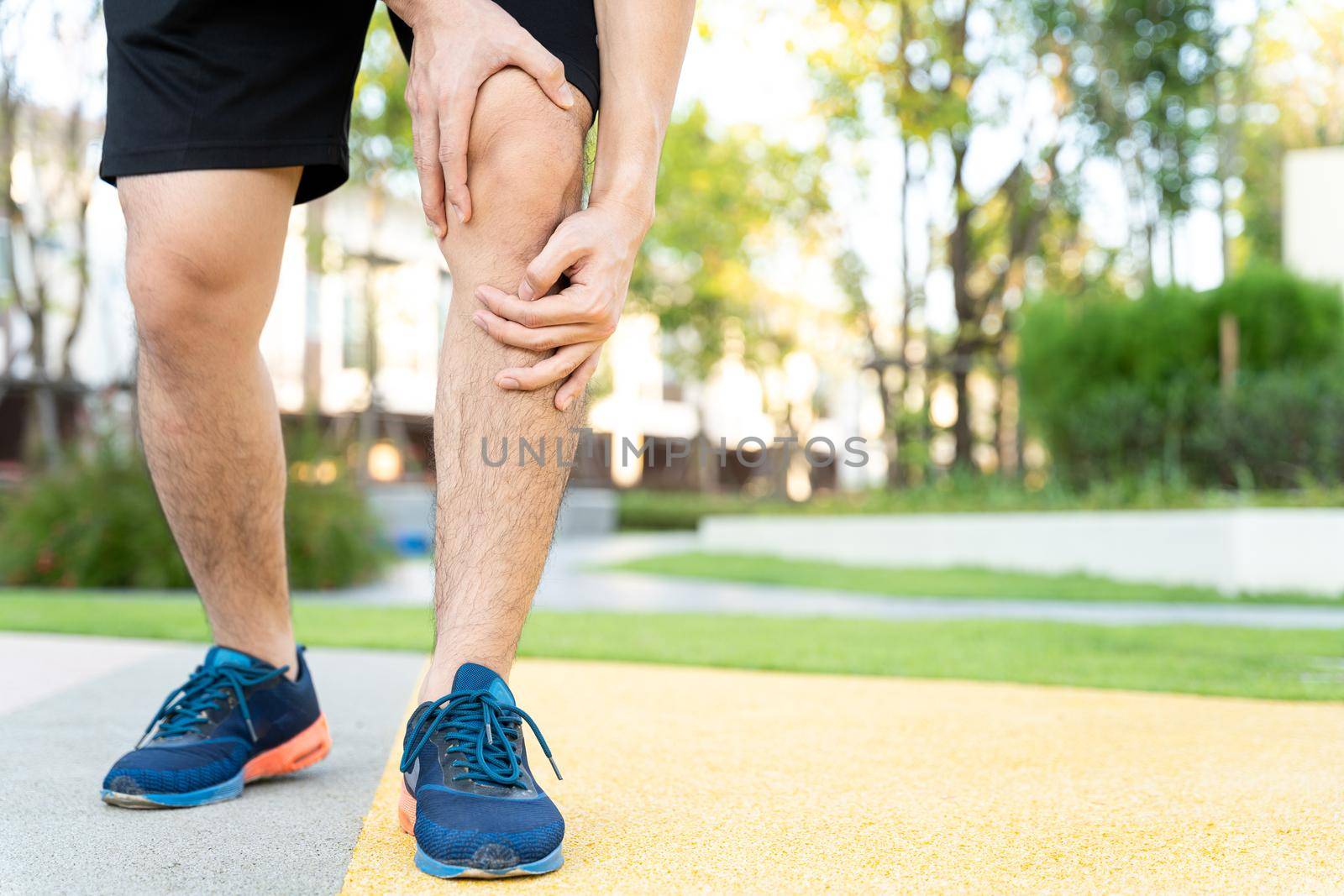 Male runner athlete leg injury and pain. Hands grab painful knee while running in the park.