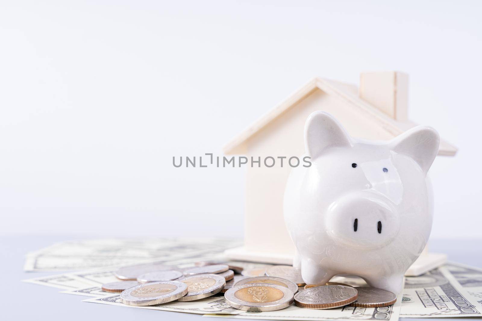 Piggy bank with wooden house and a pile of money coins and paper isolated grey background. Property investment and house mortgage financial concept. by mikesaran