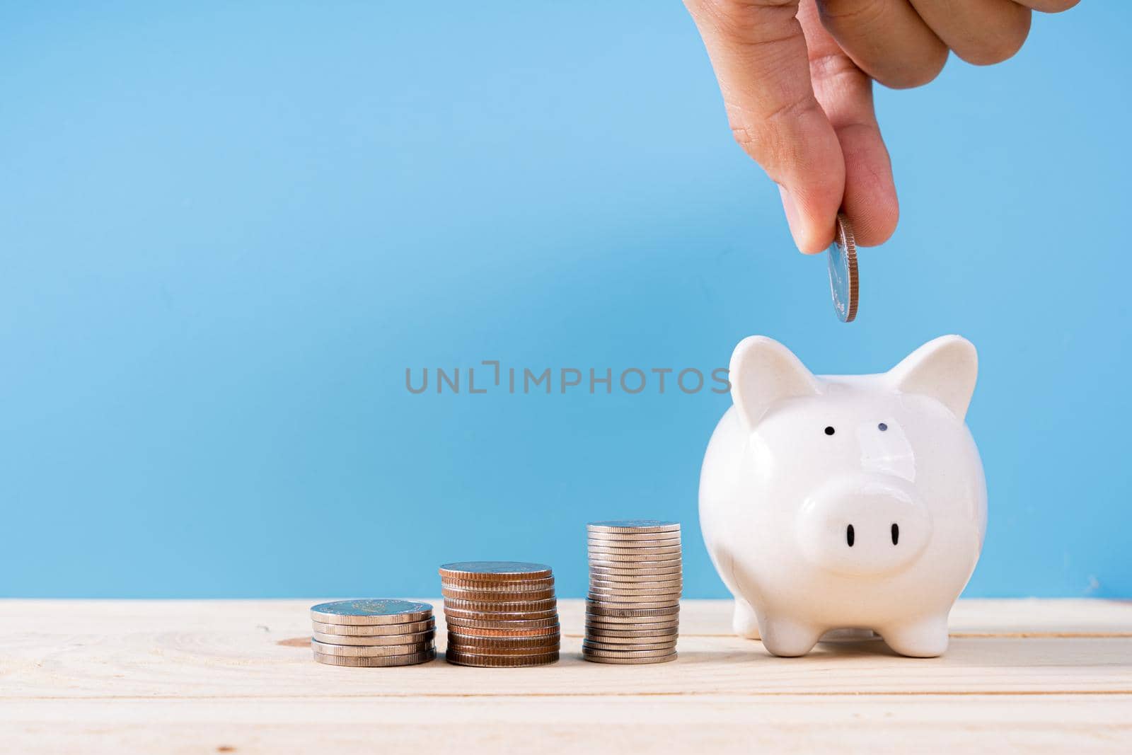 Hand putting money coin into piggy bank with stack coins isolated blue background. Saving money and investment concept by mikesaran