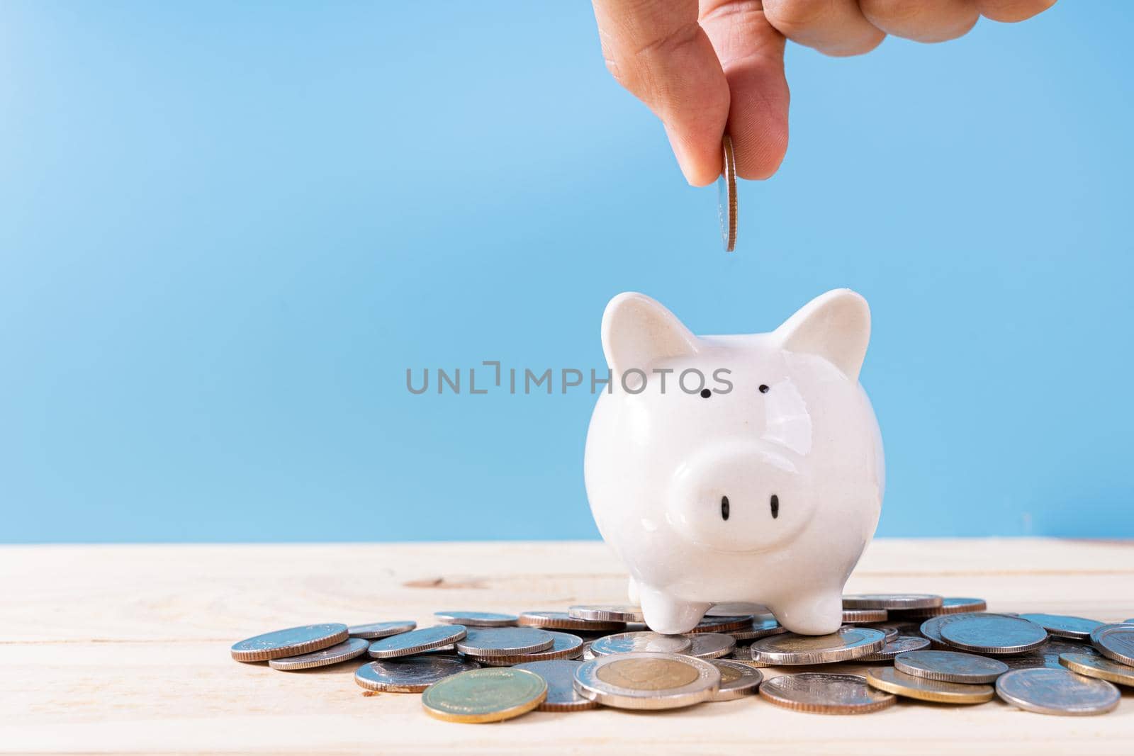 Hand putting money coin into piggy bank with a pile coins isolated blue background. Saving money and investment concept.