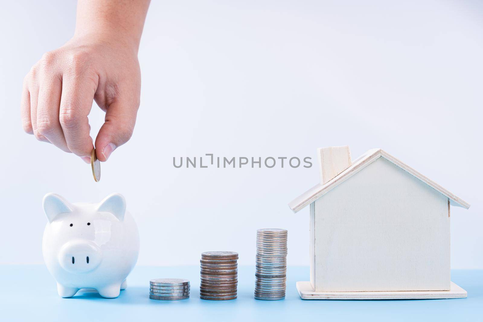 Hand putting money coin into piggy bank with wooden house and stack coins isolated grey background. Property investment and house mortgage financial concept.