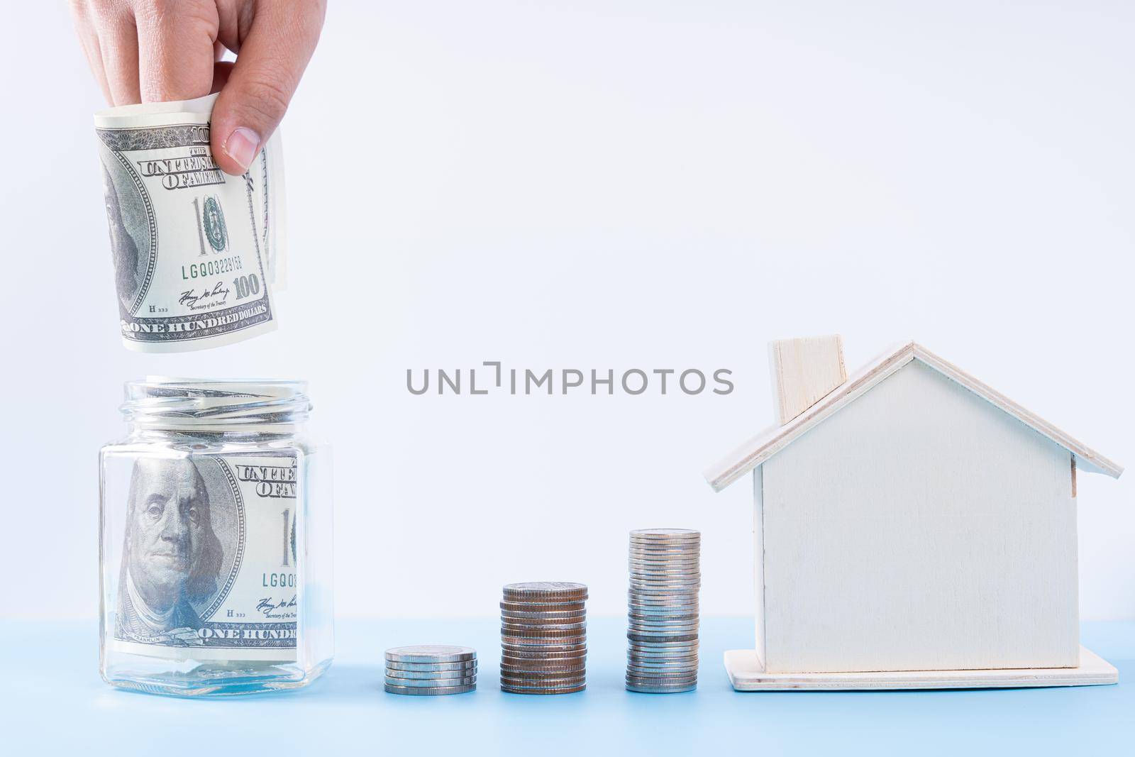 Hand putting money paper into jar with wooden house and stack coins isolated grey background. Property investment and house mortgage financial concept.