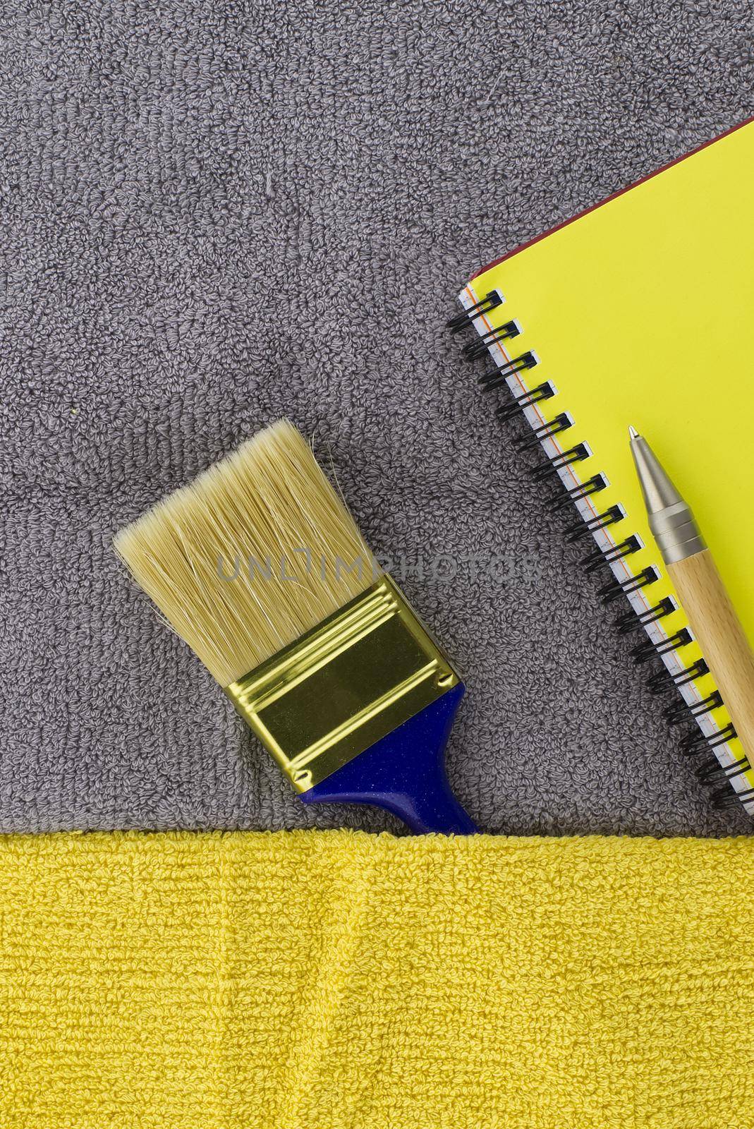 Colorful yellow and grey themed still life with clean paintbrush and yellow wire bound notepad on a two tone grey and yellow textile background
