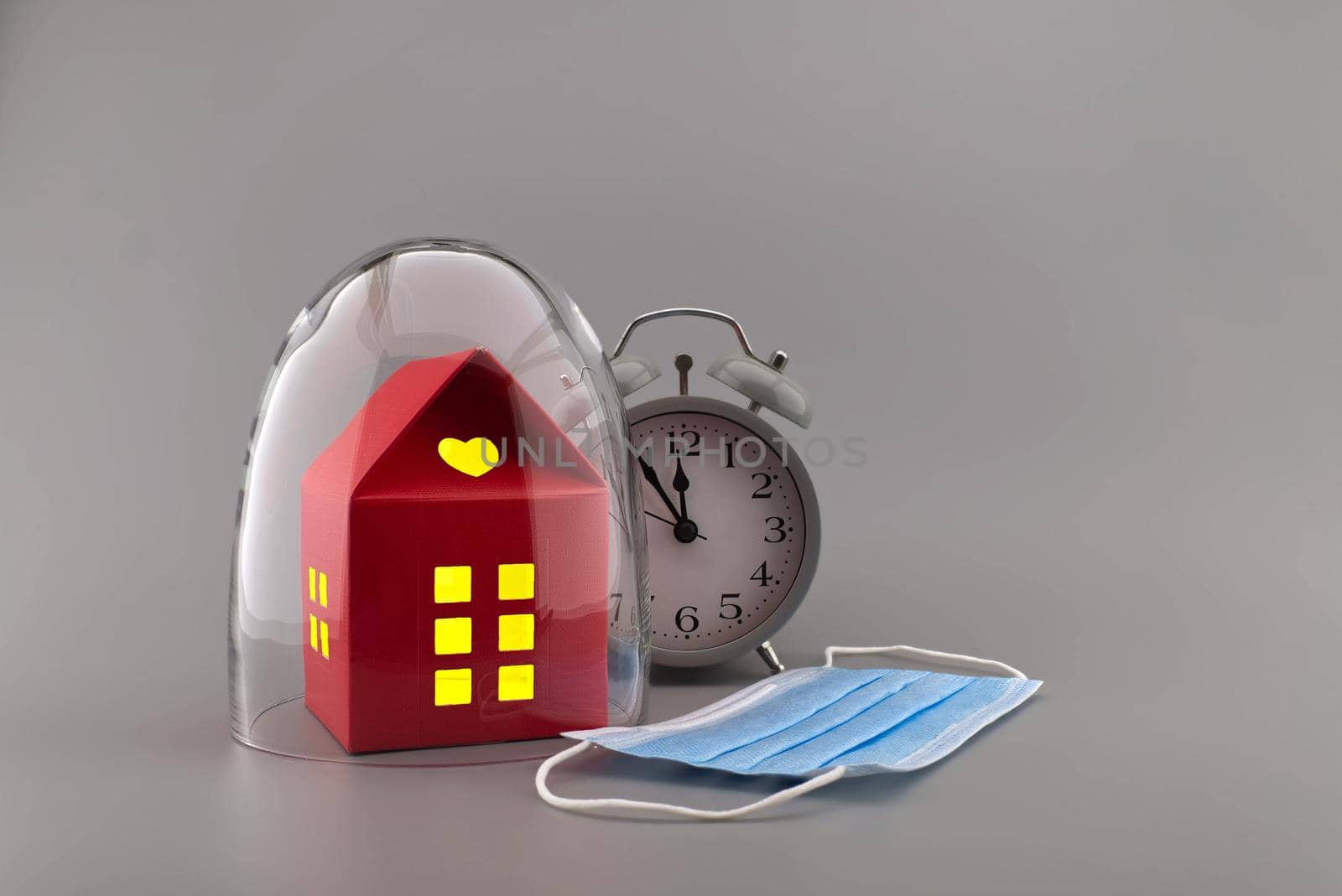 Red building under protective shield as a concept of covid pandemic and self-isolation, with the alarm clock and fave mask outside the glass dome